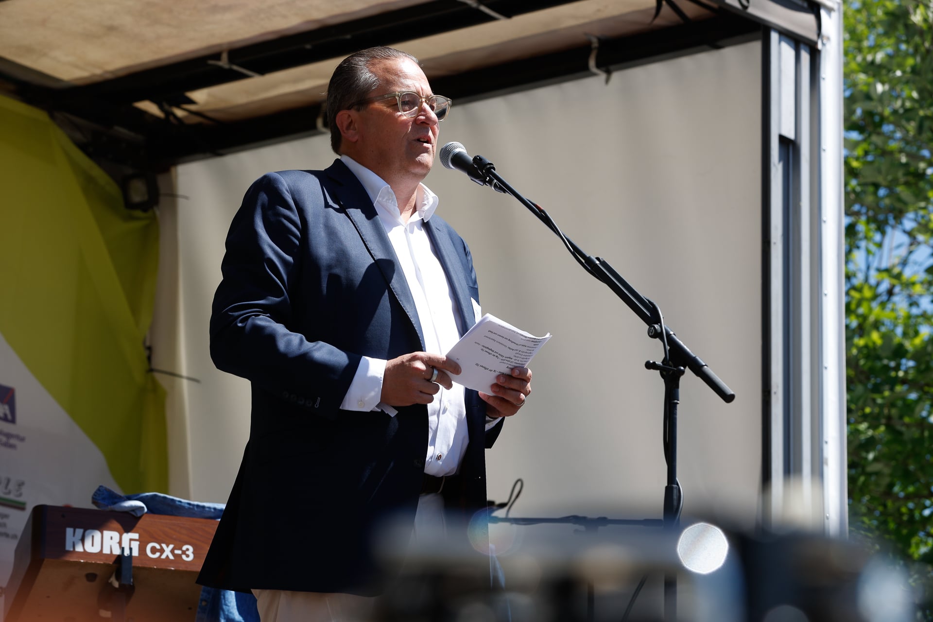 Ein Mann im Anzug und mit Brille spricht auf einer Freiluftbühne in ein Mikrofon und hält dabei einige Blätter in den Händen. In der Nähe steht eine Musikanlage, und Sonnenlicht fällt auf ihn.