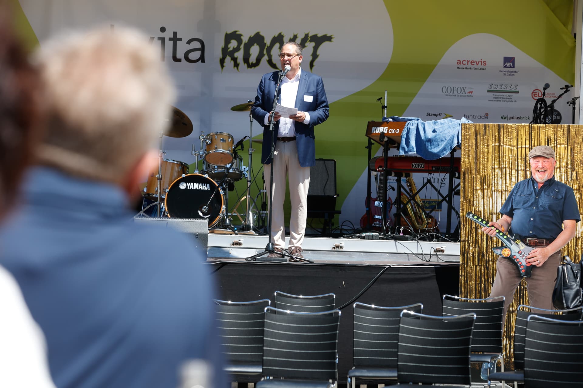 Ein Mann im Anzug spricht auf einer Bühne im Freien, hinter ihm stehen Musikinstrumente. Ein Publikum schaut zu, und auf der rechten Seite der Bühne steht ein großes Foto eines lächelnden Mannes mit einem Saxophon.