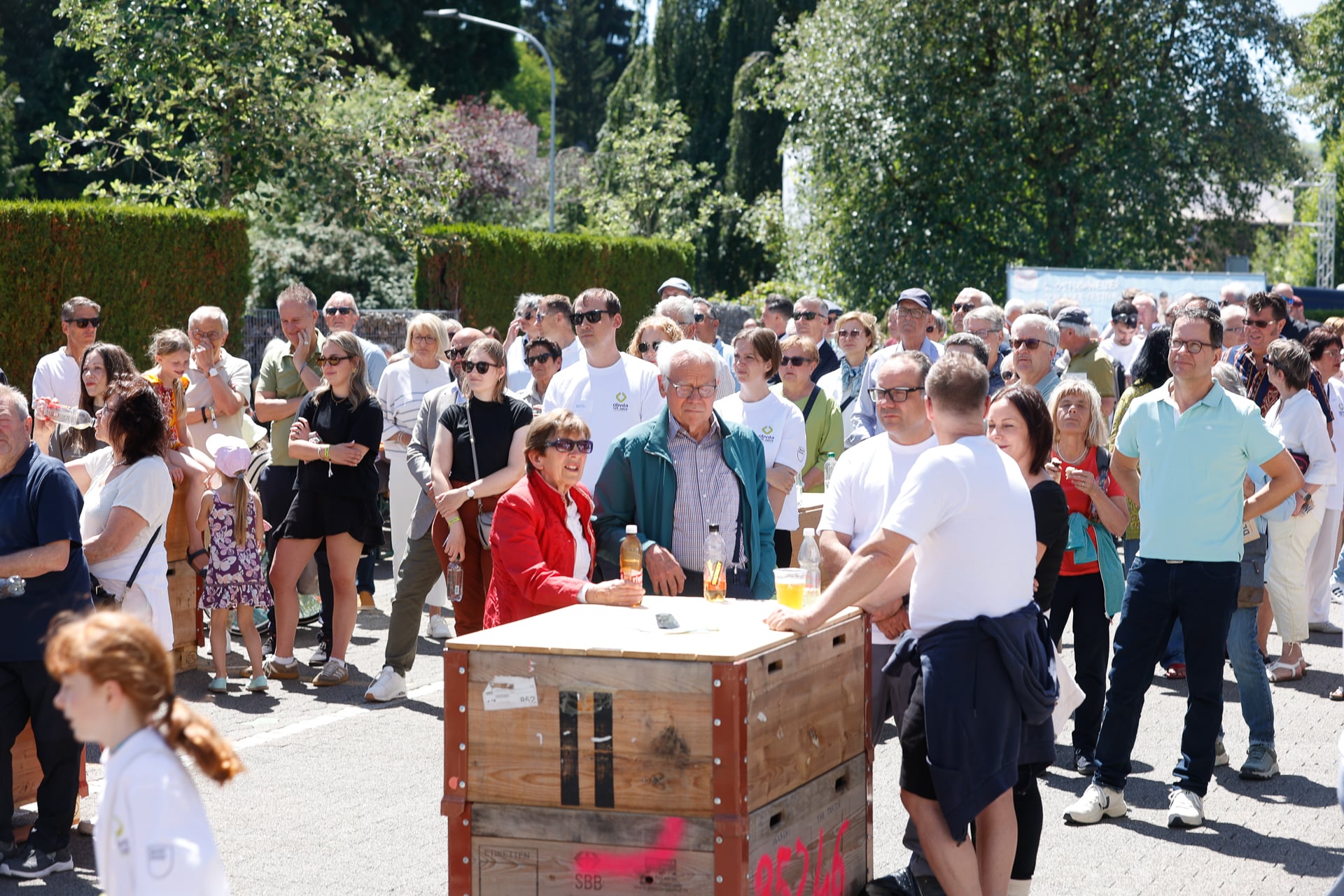 An einem sonnigen Tag versammelt sich draußen eine große Gruppe von Menschen, von denen einige Sonnenbrillen tragen, um eine Holzkiste herum. Einige von ihnen halten Getränke in der Hand, und im Hintergrund sind Bäume und Sträucher zu sehen.
