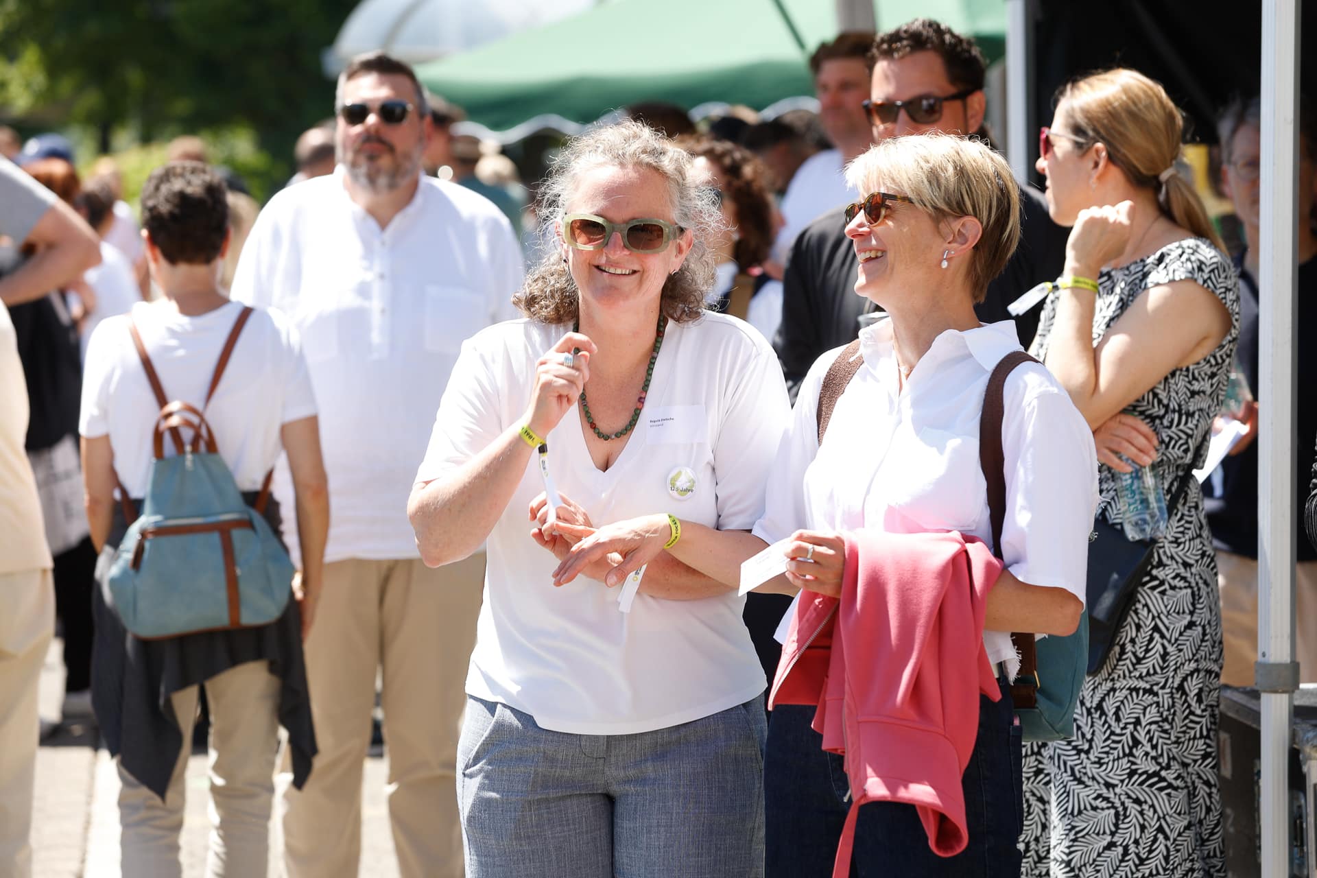 Zwei Frauen, die bei einer Veranstaltung im Freien inmitten einer Menschenmenge lächeln und sich unterhalten; beide tragen Sonnenbrillen und helle Kleidung. Auch die Menschen im Hintergrund sind leger gekleidet und genießen den sonnigen Tag.