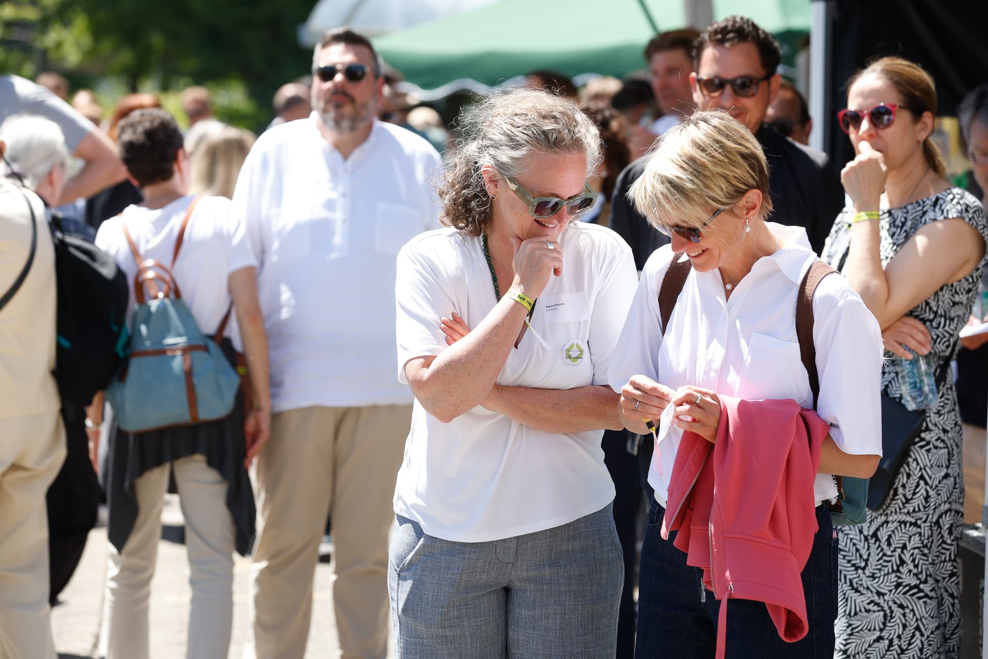 Zwei Frauen stehen dicht beieinander, lächeln und betrachten etwas, das eine von ihnen in der Hand hält, umgeben von einer Menschenmenge im Freien an einem sonnigen Tag. Eine der Frauen hält eine rosa Jacke in der Hand. Die Atmosphäre ist ungezwungen und lebhaft.
