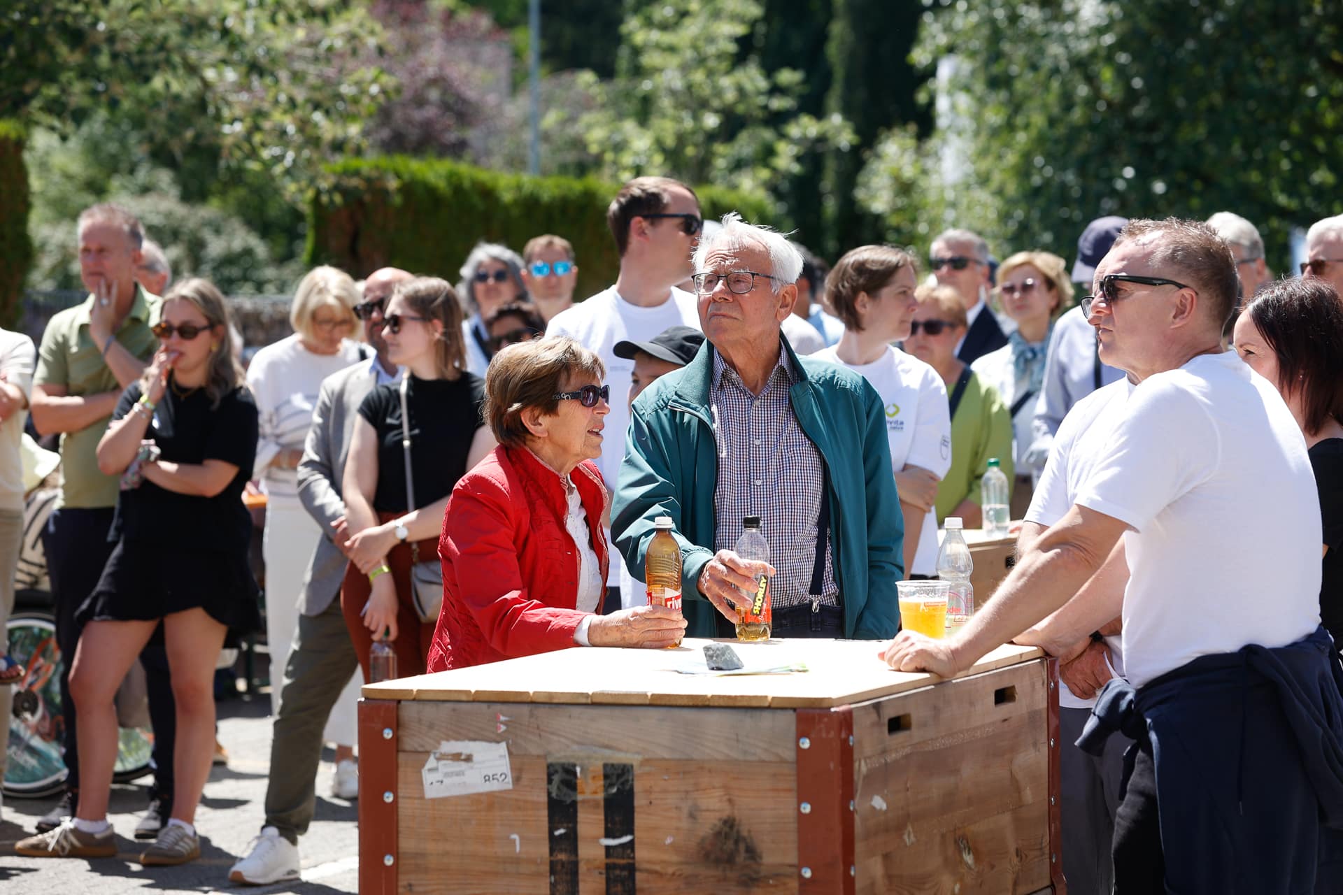 Eine Gruppe von Menschen hat sich draußen um einen Holztisch versammelt, einige halten Getränke in der Hand. Ein älterer Mann und eine ältere Frau stehen am Tisch, während andere Personen in Freizeitkleidung daneben stehen und etwas außerhalb des Bildausschnitts beobachten. Es ist sonnig und alles ist grün.