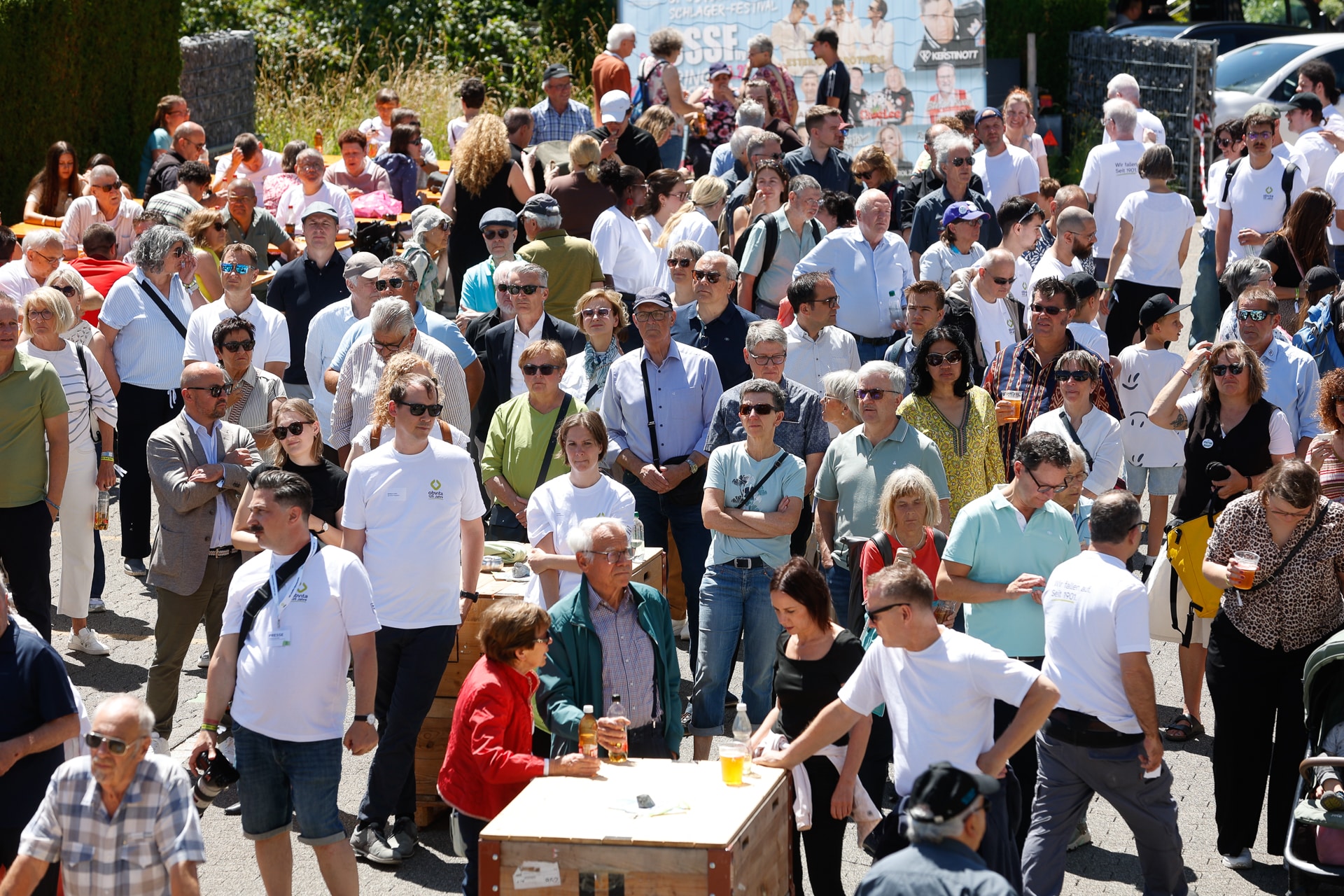 An einem sonnigen Tag versammelt sich draußen eine große Menschenmenge, von der viele eine Sonnenbrille tragen. Einige stehen, andere sitzen, und ein paar halten Getränke in der Hand. Im Hintergrund sind grüne Bäume und Plakate zu sehen.