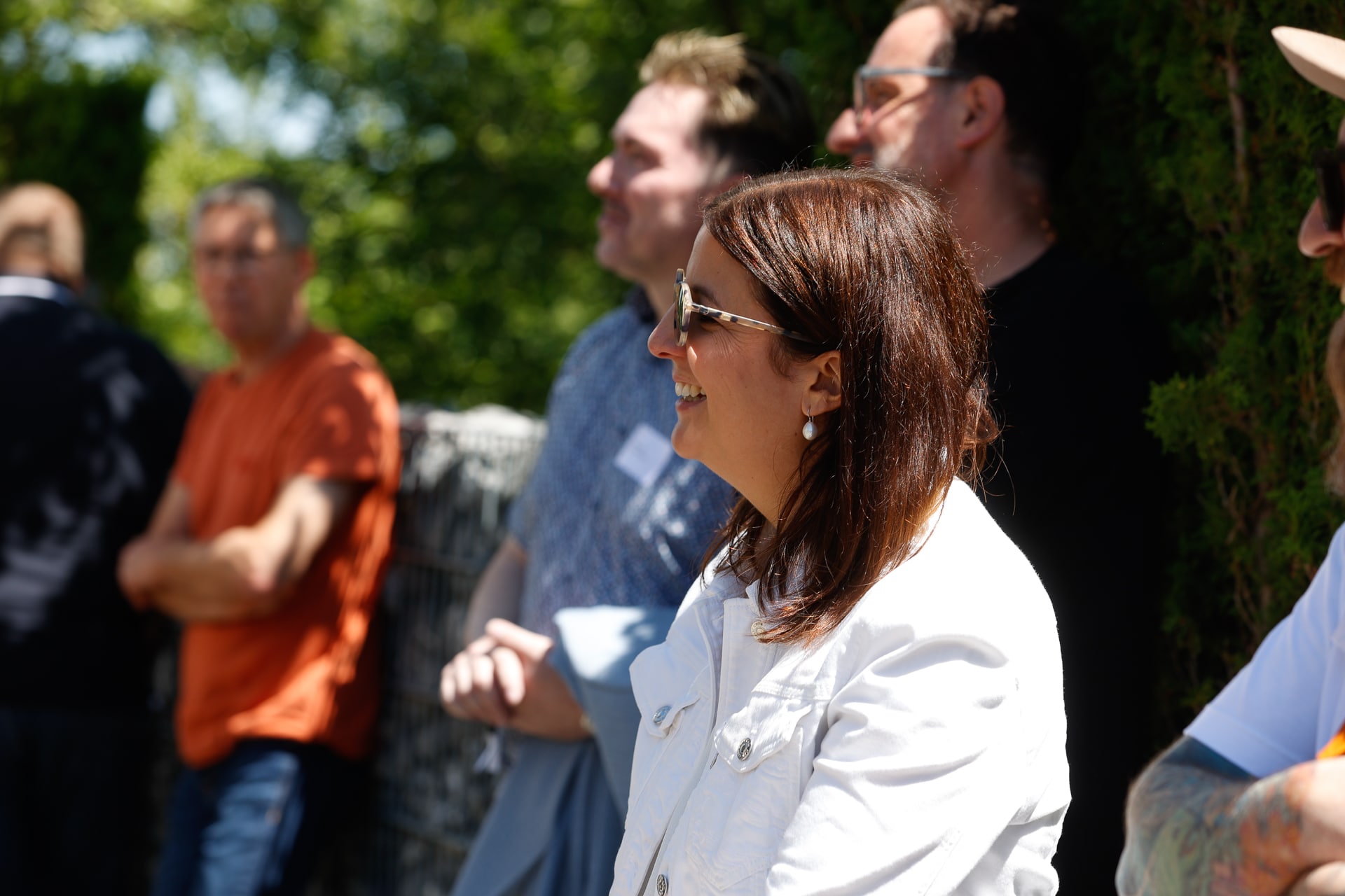 Eine Frau mit Sonnenbrille und weißer Jacke lächelt draußen, umgeben von mehreren Menschen, die in der Sonne stehen, im Hintergrund sind grüne Bäume zu sehen.