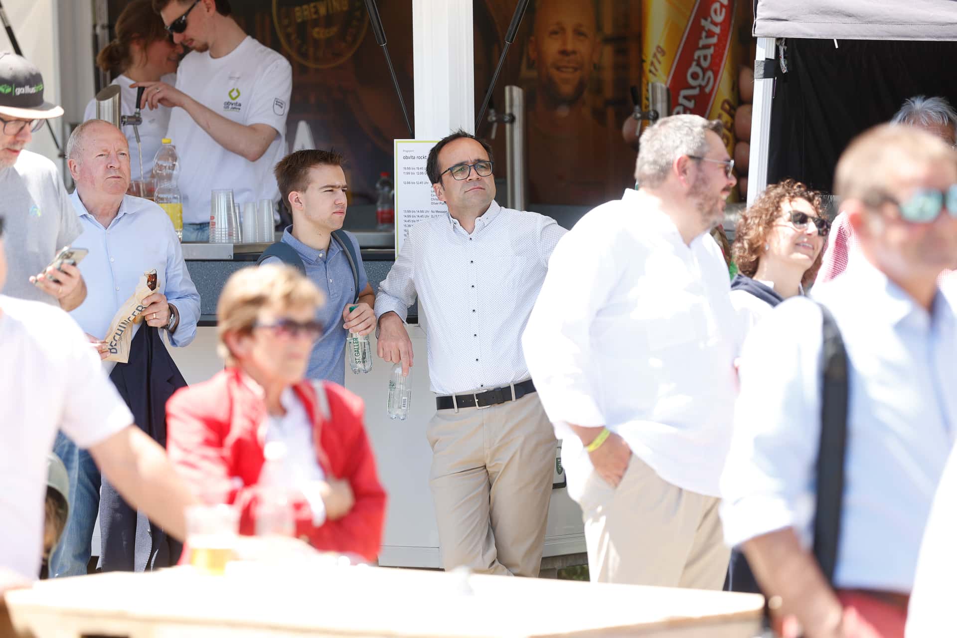 Eine Gruppe von Menschen, von denen die meisten Business- oder Smart-Casual-Kleidung tragen, steht draußen im Sonnenlicht; einige halten Getränke in der Hand und blicken aufmerksam in dieselbe Richtung. Die Szene scheint bei einer Veranstaltung oder einem Treffen aufgenommen worden zu sein.
