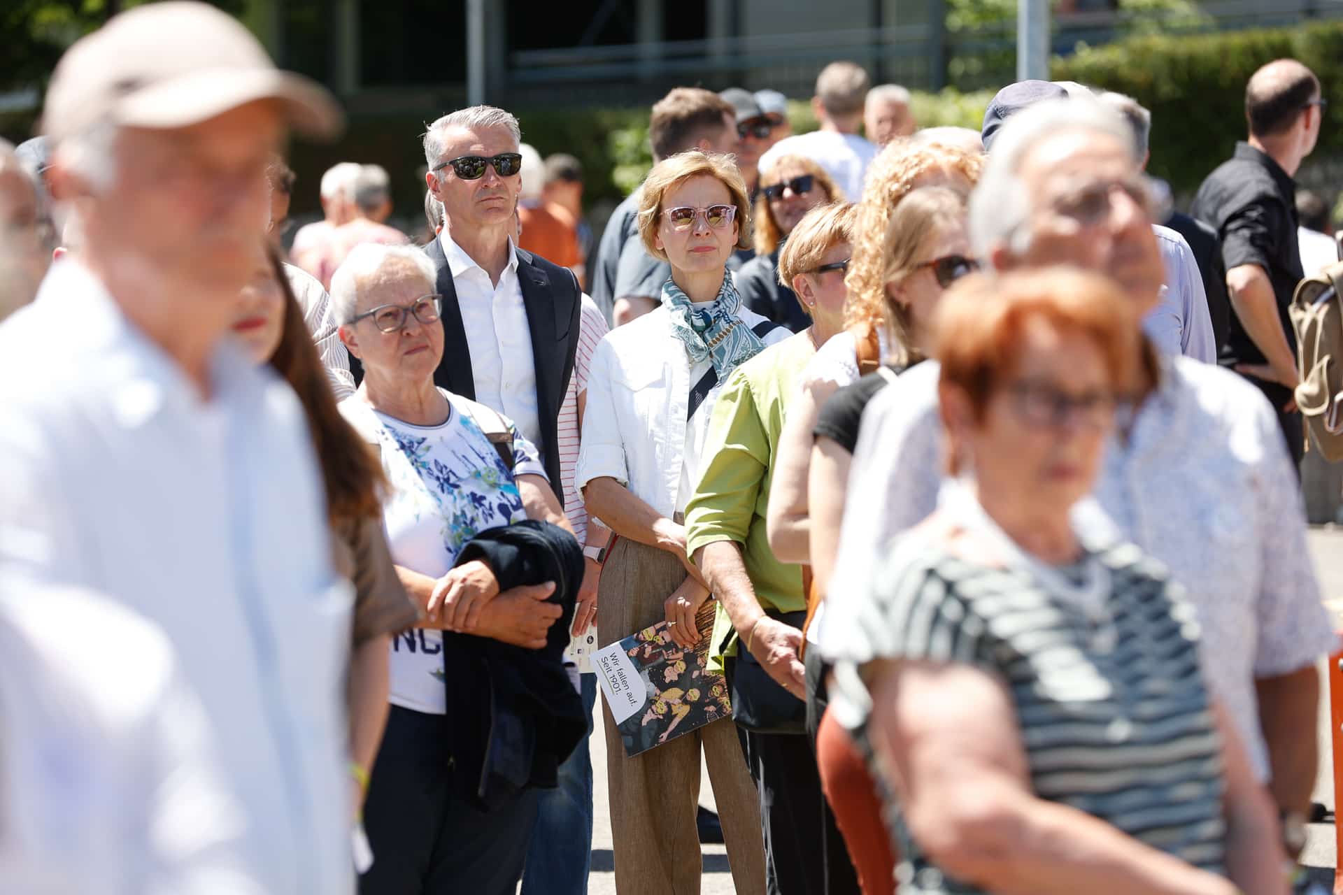 Eine Gruppe von Menschen steht draußen im Sonnenlicht und hört aufmerksam zu. Einige tragen Sonnenbrillen und leichte Kleidung. Im Mittelpunkt steht eine Frau in der Mitte, die eine Broschüre in der Hand hält, während andere Personen im Vordergrund leicht unscharf erscheinen.