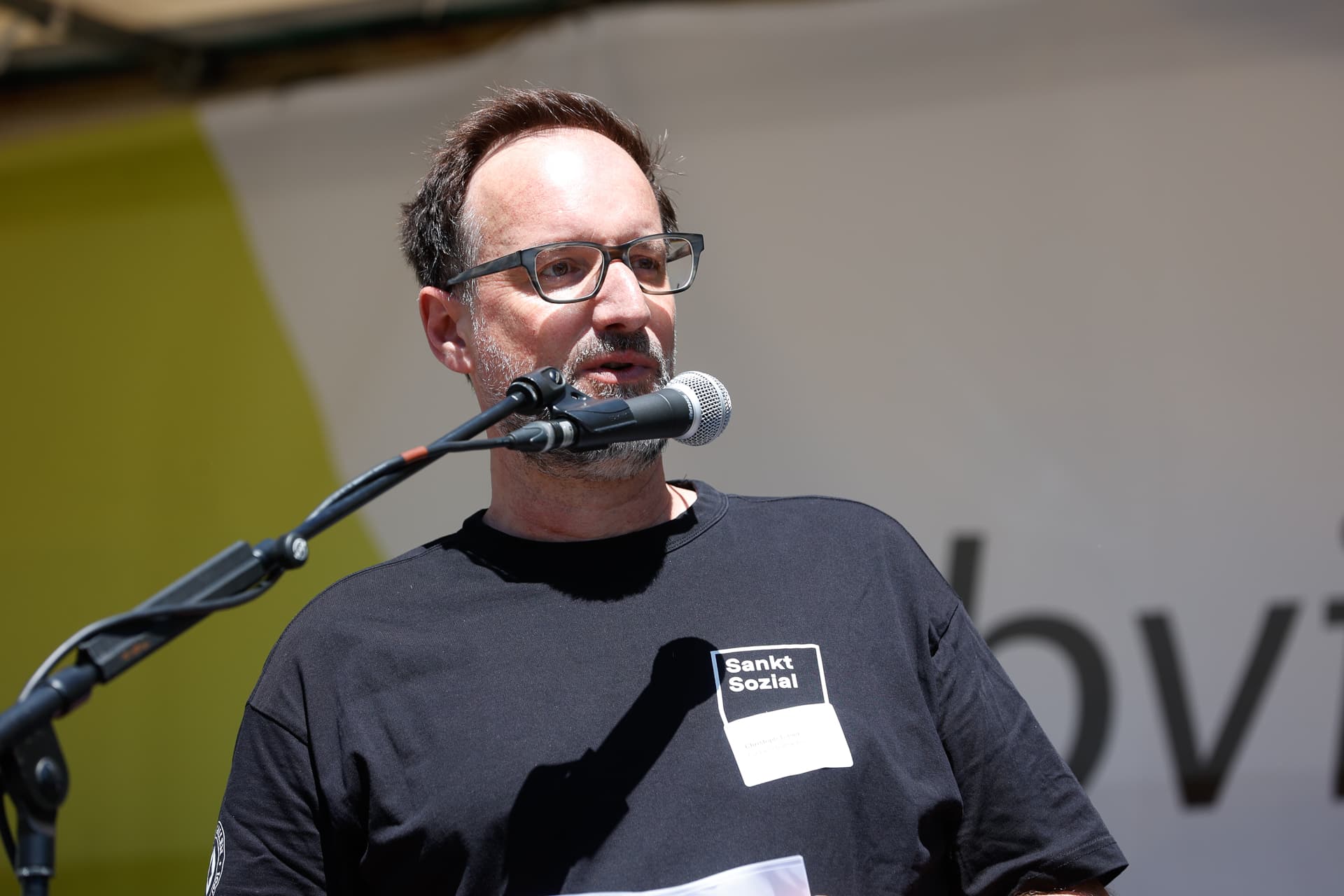 Ein Mann mit Brille und Bart, der ein schwarzes „Sankt Sozial“-T-Shirt trägt, spricht bei einer Veranstaltung im Freien in ein Mikrofon. Er hält einige Blätter in der Hand und steht vor einem unscharfen Hintergrund.