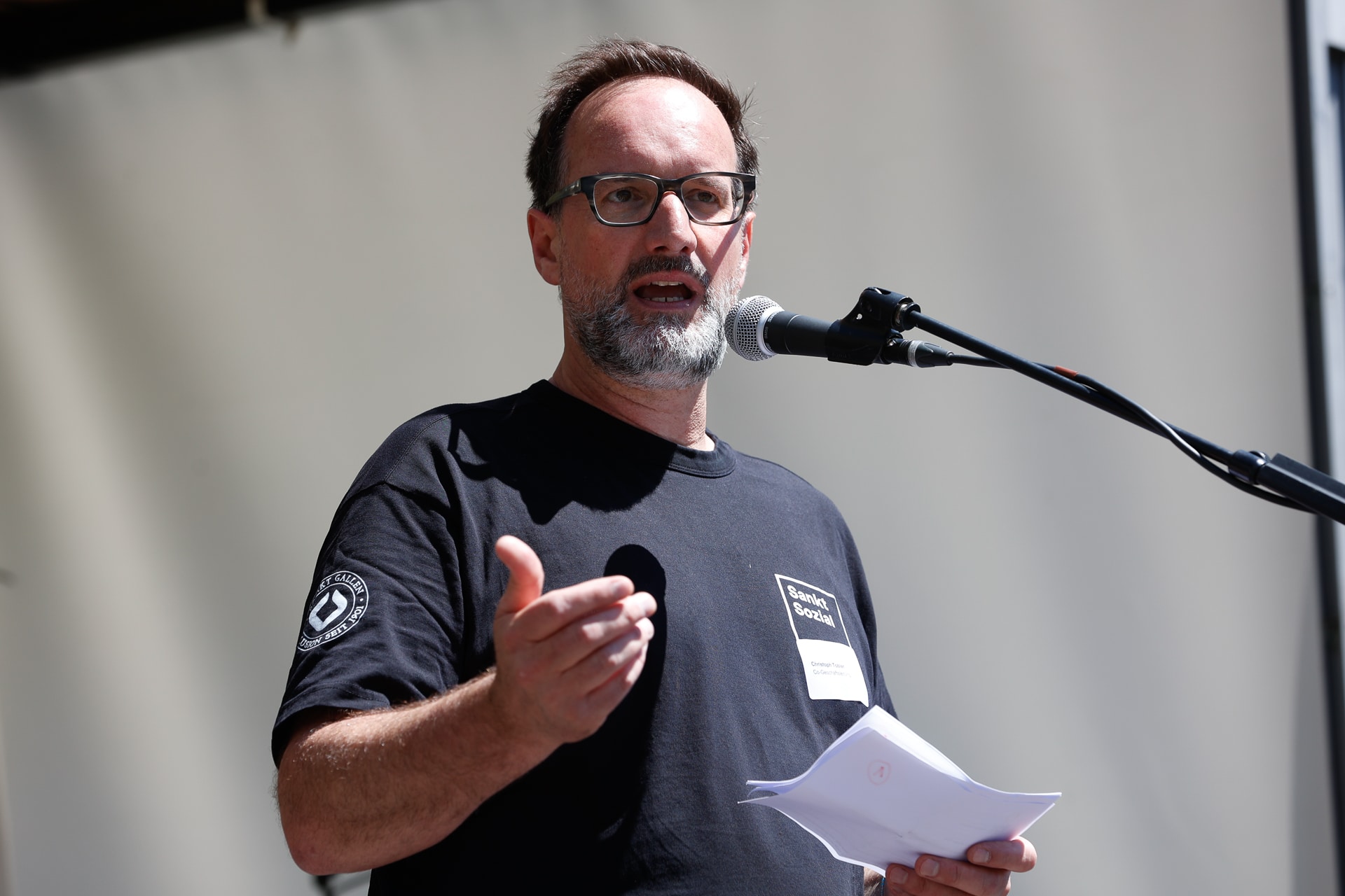 Ein Mann mit Brille und Bart spricht in ein Mikrofon, während er einige Blätter in der Hand hält. Er trägt ein schwarzes T-Shirt und steht vor einem schlichten Hintergrund.
