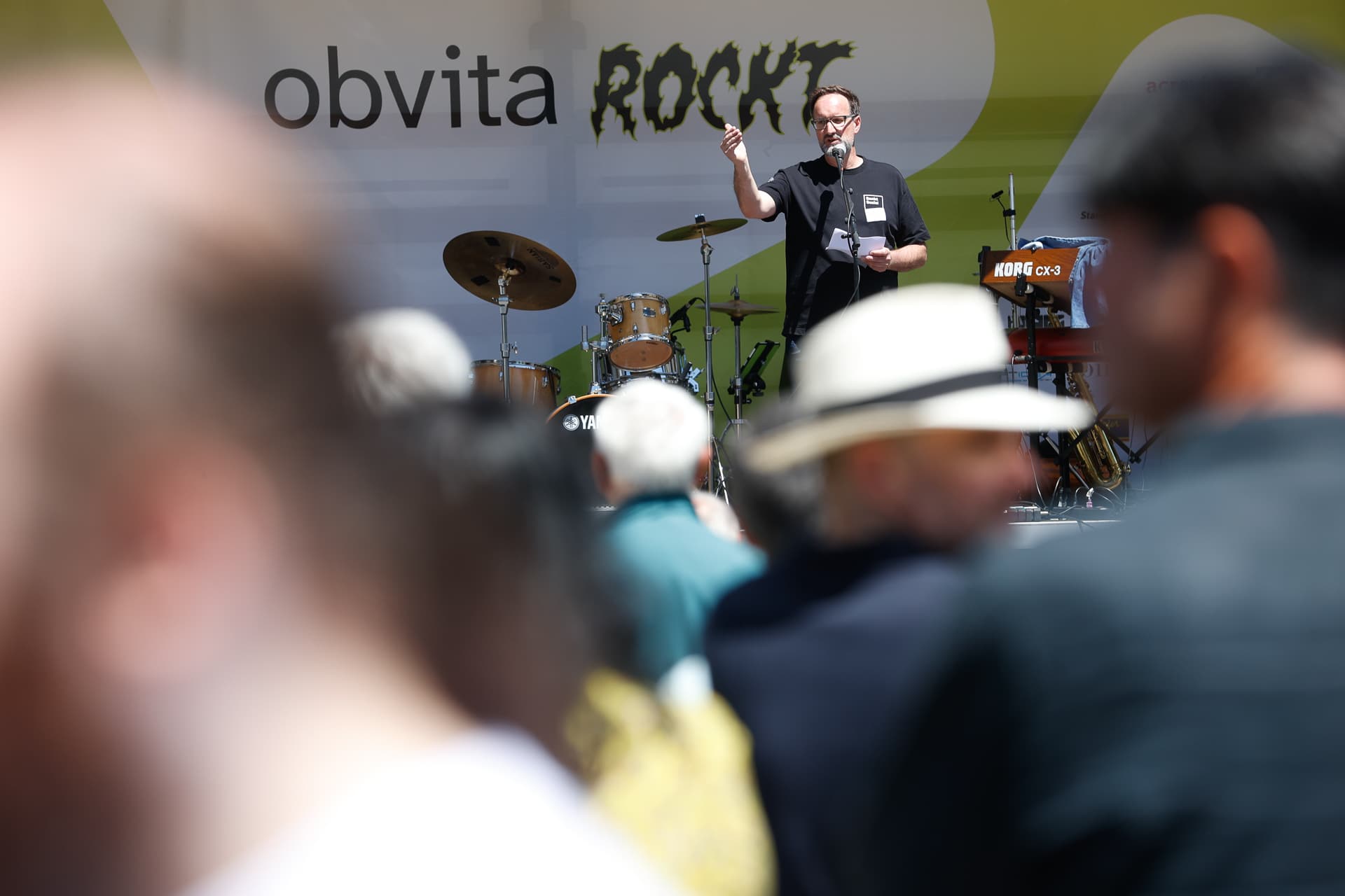 Eine Person spricht auf einer Außenbühne mit Schlagzeug und Keyboard vor einem Publikum in ein Mikrofon. Auf dem Bühnenhintergrund steht „obvita ROCKT“. Das Foto wurde über die Schultern der Zuschauer hinweg aufgenommen.