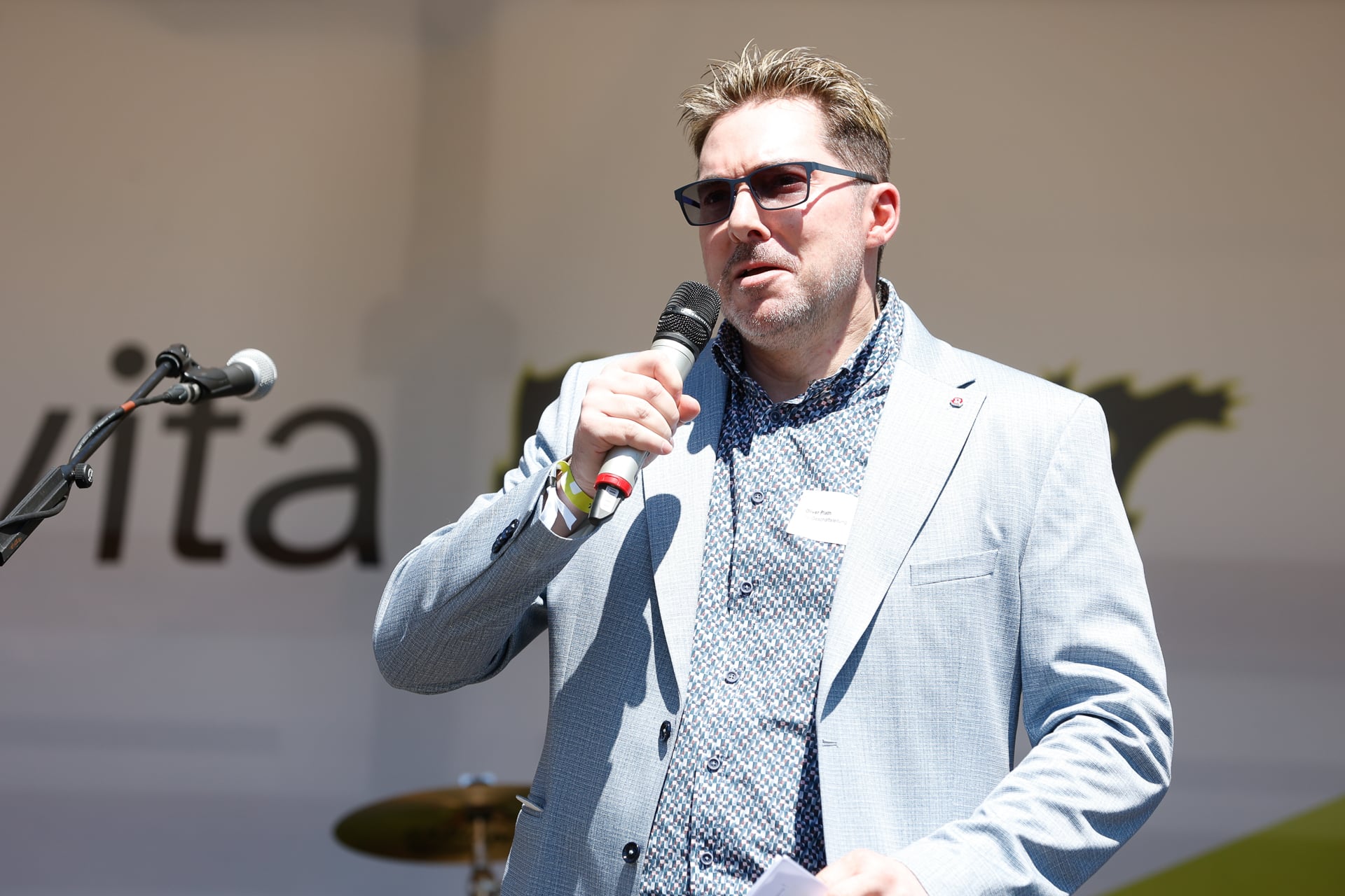 Ein Mann mit Sonnenbrille und hellgrauem Blazer hält ein Mikrofon in der Hand, während er auf einer Bühne im Freien spricht. Der Hintergrund ist unscharf, teilweise sind Textzeilen und ein Schlagzeug zu erkennen.