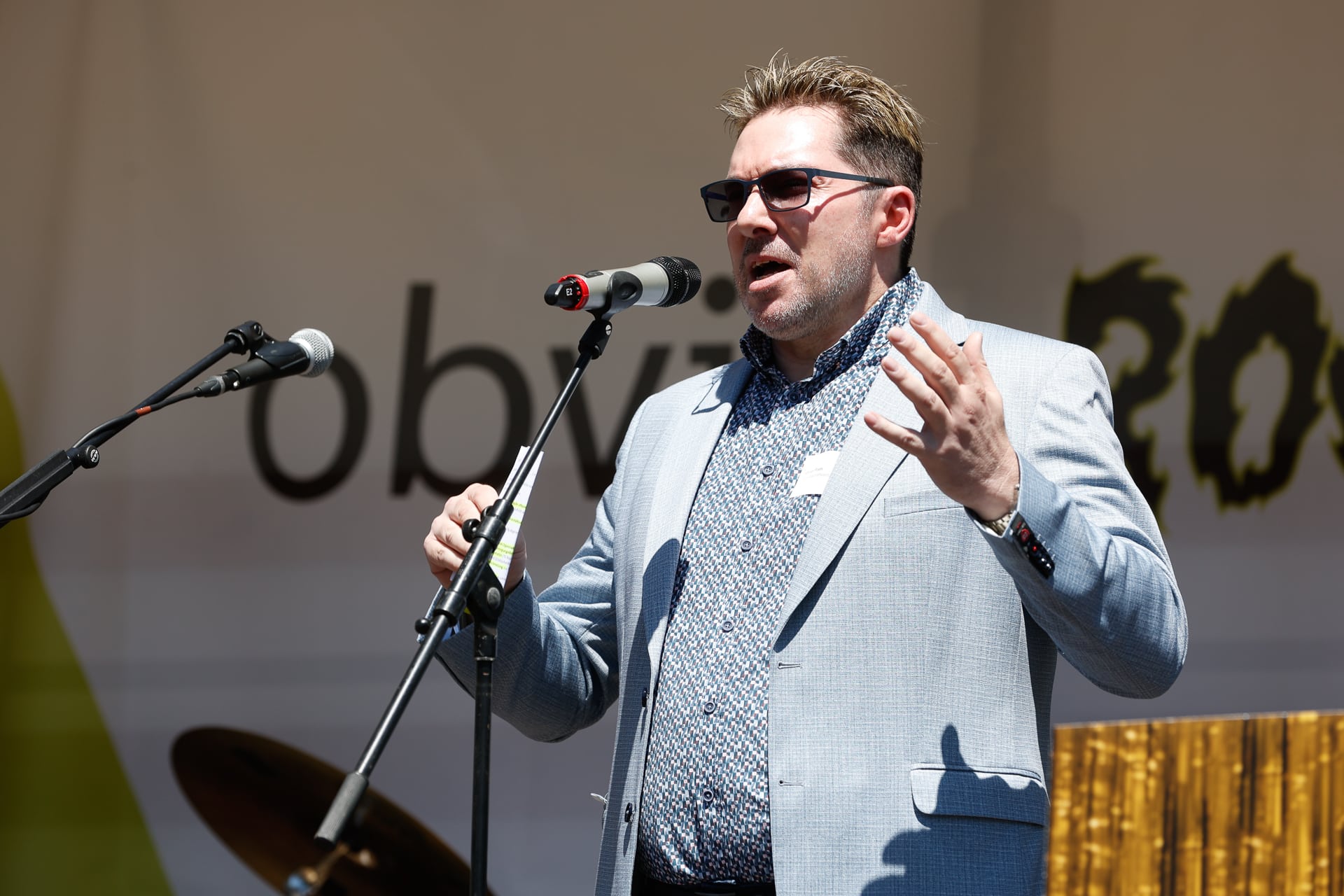 Ein Mann mit Sonnenbrille und hellblauem Blazer spricht auf einer Freiluftbühne leidenschaftlich in ein Mikrofon und gestikuliert dabei mit einer Hand.