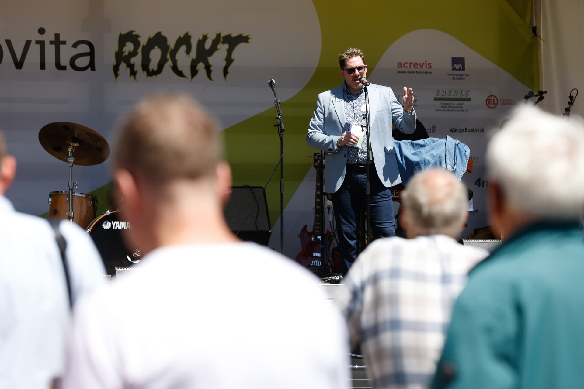Ein Mann im Anzug spricht mit einem Mikrofon auf einer Bühne im Freien und blickt dabei ins Publikum. Im Hintergrund sind Musikinstrumente und ein „ROCKT“-Schild zu sehen. Im Vordergrund schauen mehrere Personen zu.