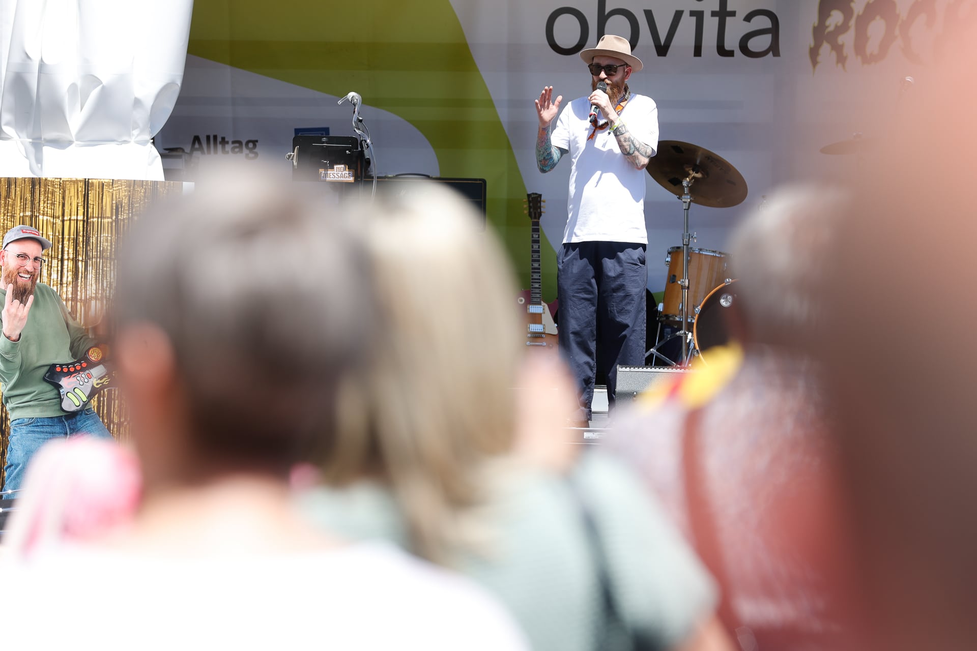 Ein Mann mit Hut und Sonnenbrille spricht oder singt auf einer Freiluftbühne in ein Mikrofon, während ein anderer Mann in der Nähe kniet und Gitarre spielt. Im Vordergrund ist eine verschwommene Menschenmenge zu sehen, die der Darbietung zusieht.