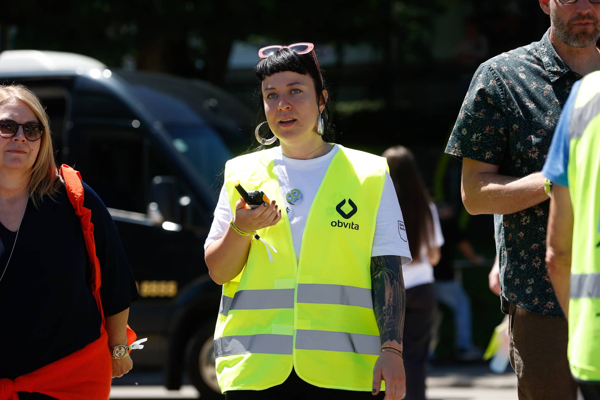 Eine Frau, die eine neongelbe Sicherheitsweste trägt und ein Walkie-Talkie in der Hand hält, steht zusammen mit anderen Personen im Freien. Auf der Weste ist das „obvita“-Logo zu sehen. Im Hintergrund sind ein Lkw und Bäume zu erkennen.