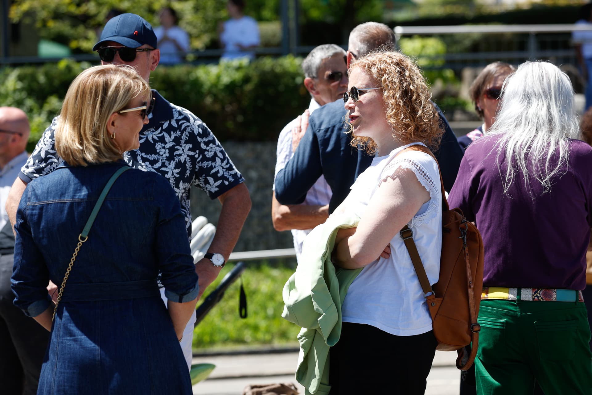 Eine Gruppe Erwachsener steht im Freien und unterhält sich im Sonnenlicht. Einige tragen Sonnenbrillen. Eine Frau mit lockigem Haar und einem weißen Oberteil spricht mit einer Frau in einer Jeansjacke. Im Hintergrund sind Grünflächen und Menschen zu sehen.