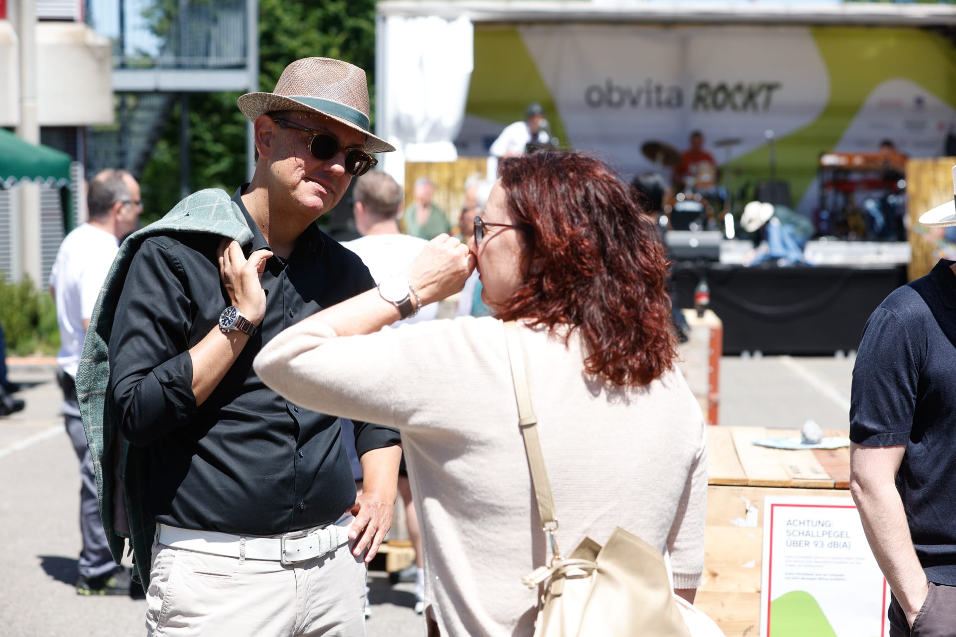 Zwei Menschen unterhalten sich an einem sonnigen Tag bei einer Veranstaltung im Freien. Ein Mann mit Sonnenbrille hält sich eine Jacke über die Schulter, während ihm eine Frau mit roten Haaren gegenübersteht. Im Hintergrund sind eine Bühne und weitere Personen zu sehen.