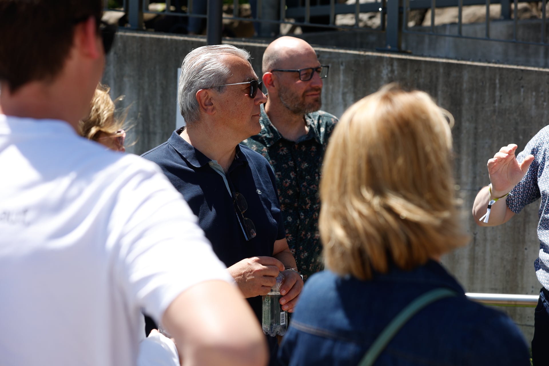 Eine Gruppe von Menschen steht draußen im Sonnenlicht und unterhält sich. Ein Mann mit Sonnenbrille und einem marineblauen Poloshirt hält eine Dose in der Hand, während andere mit dem Rücken zur Kamera stehen oder nur teilweise zu sehen sind. Im Hintergrund sind eine Betonmauer und Metallgeländer zu erkennen.