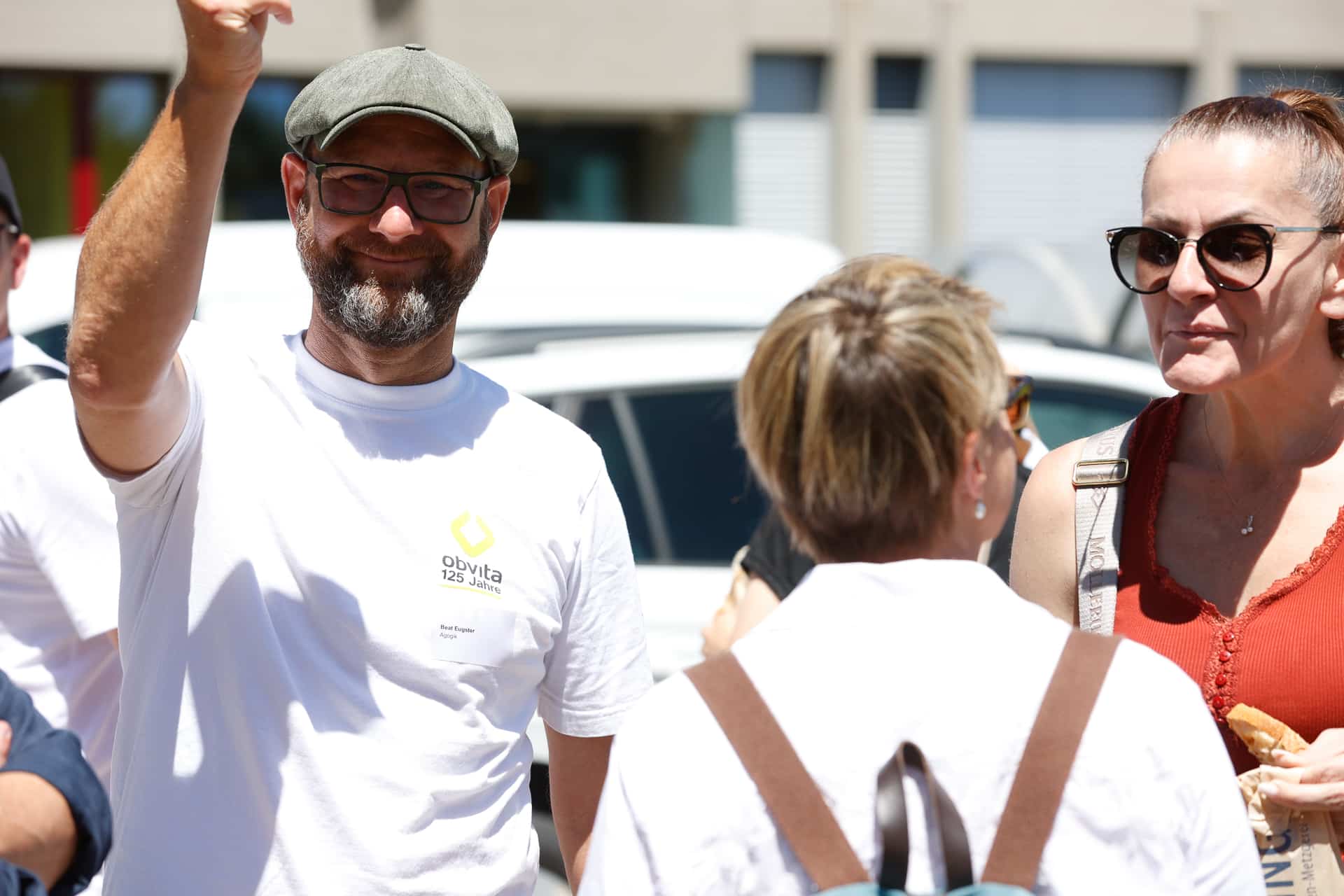Ein Mann mit Brille, Mütze und weißem T-Shirt steht an einem sonnigen Tag draußen mit zwei Frauen im Gespräch, lächelt und winkt.