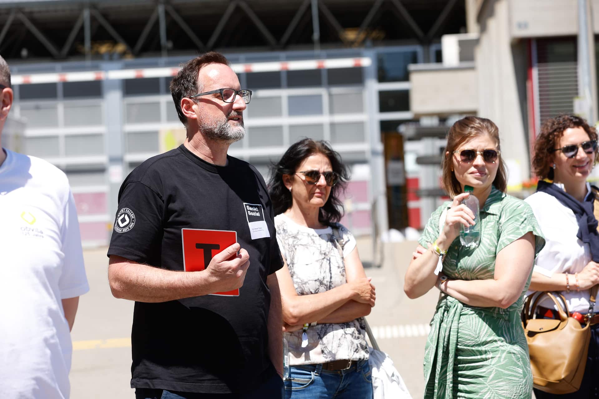 Eine Gruppe von Menschen steht an einem sonnigen Tag im Freien; ein Mann in einem schwarzen Hemd hält ein rotes Schild in der Hand. Die anderen sind leger gekleidet, einige tragen Sonnenbrillen, und im Hintergrund ist ein Gebäude zu sehen.