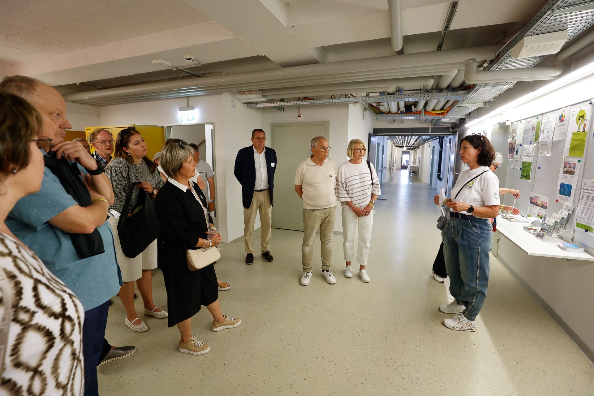 Eine Gruppe von Menschen steht in einem Flur und hört einer Frau zu, die einen Vortrag hält, während hinter ihr Plakate und Schautafeln an der Wand hängen. Die Szene scheint in einem Innenraum stattzufinden, wahrscheinlich in einer Bildungs- oder Forschungseinrichtung.