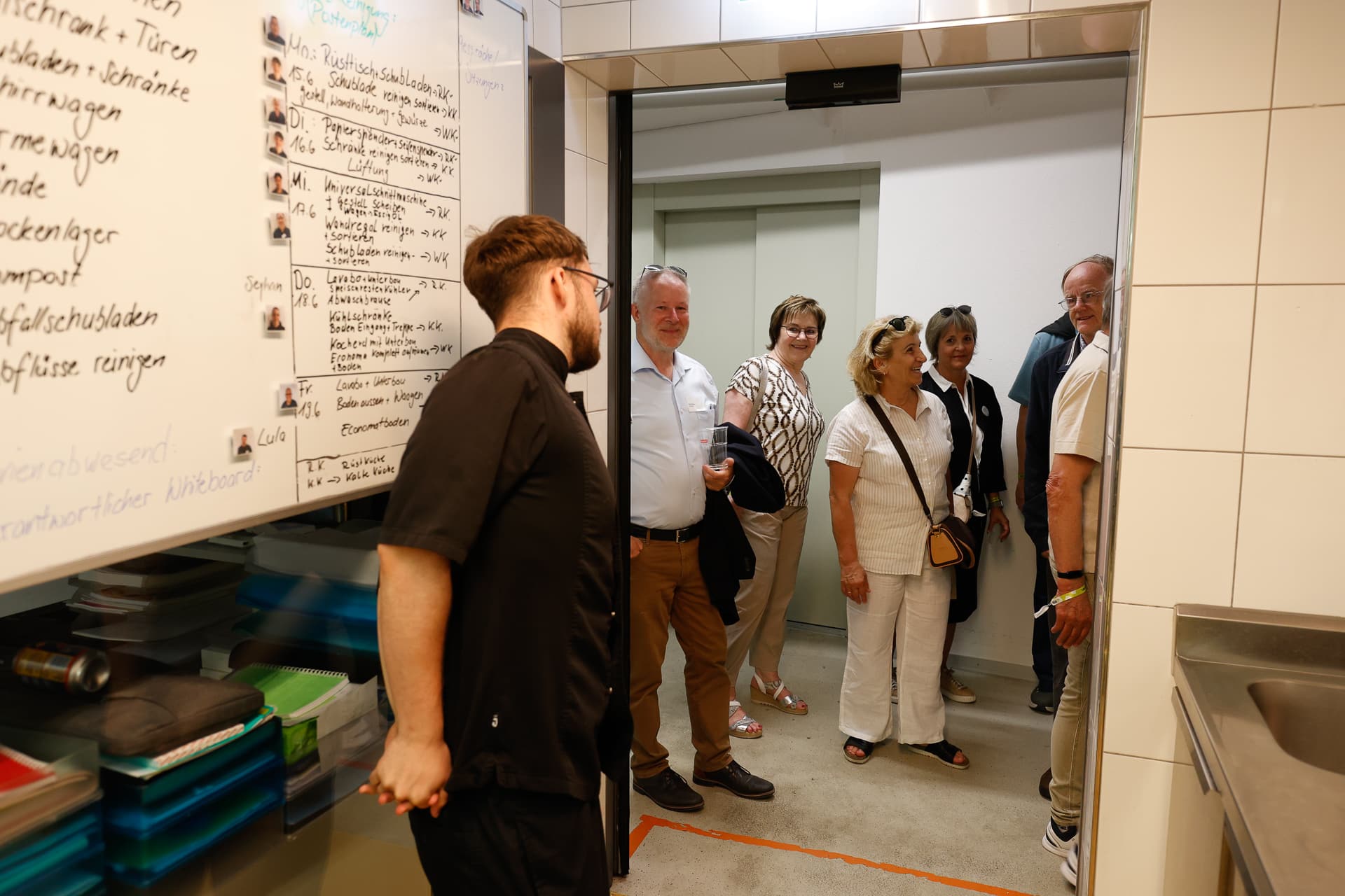 Eine Gruppe von sechs Erwachsenen steht in einem Türrahmen, lächelt und unterhält sich mit einem Mann in schwarzer Kleidung, der sich in einem mit Fliesen ausgekleideten Raum befindet, an dessen Wand eine mit handschriftlichen Notizen bedeckte Tafel hängt.