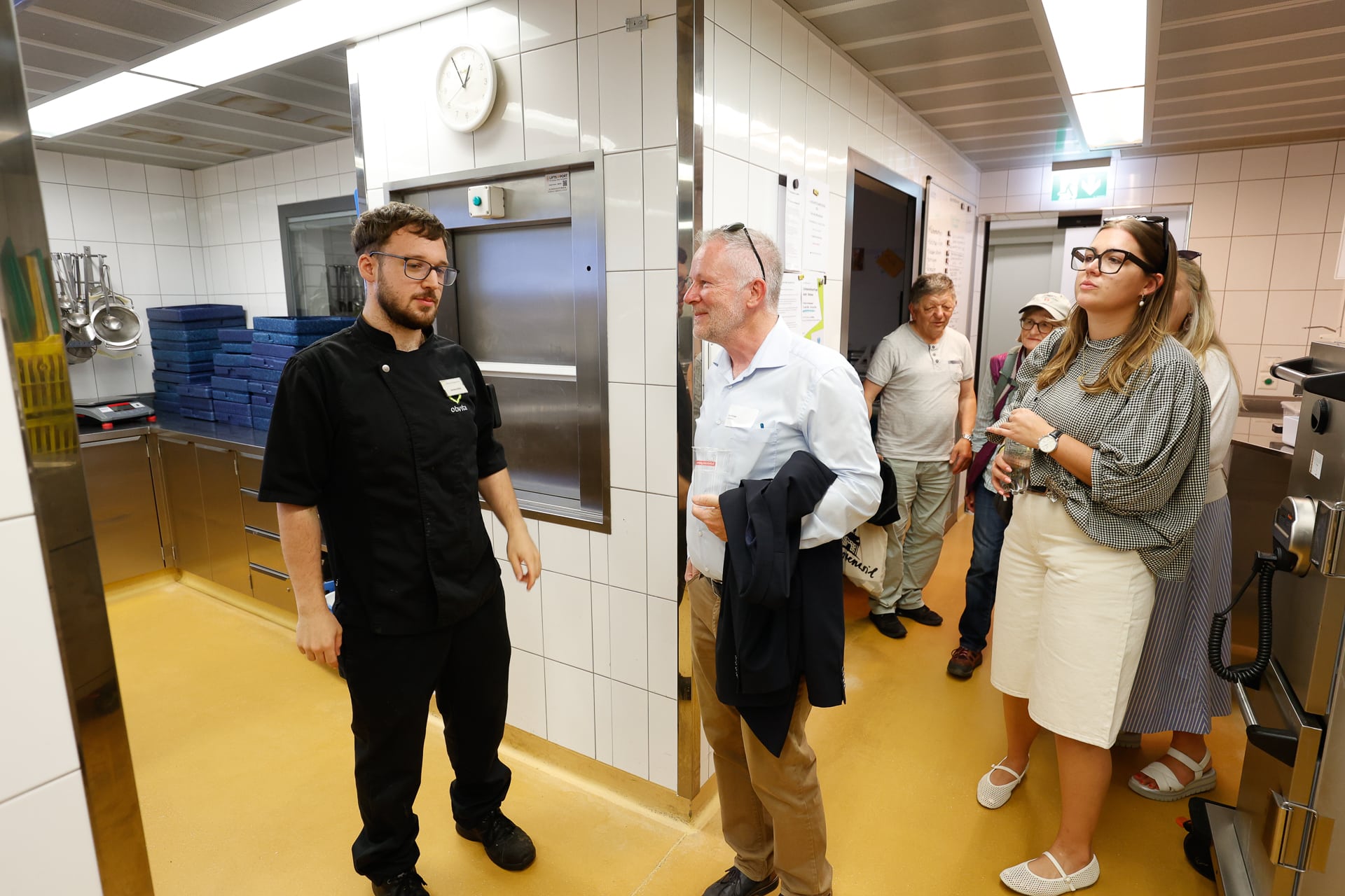 Eine Gruppe von Menschen, darunter ein Mann in einer schwarzen Kochjacke, steht in einer sauberen, modernen Küche mit weiß gefliesten Wänden, gelbem Boden und an der Wand hängenden Metallutensilien und unterhält sich.