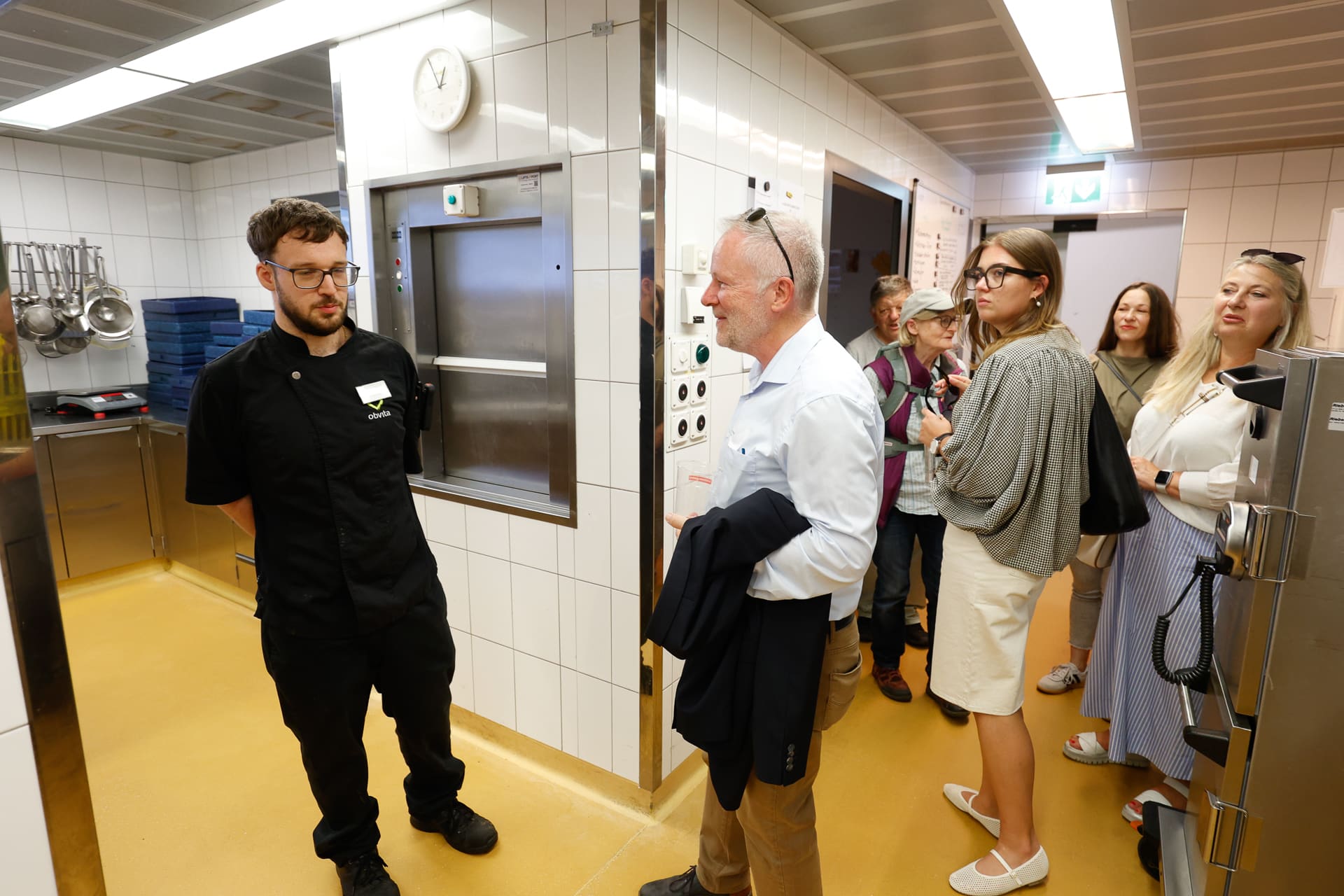 Eine Gruppe von Menschen, darunter ein Mann in einer schwarzen Kochuniform, steht in einer weiß gefliesten Küche und unterhält sich. Einige hören aufmerksam zu, während andere sich im Raum umsehen. An der Wand hängen Kochutensilien.