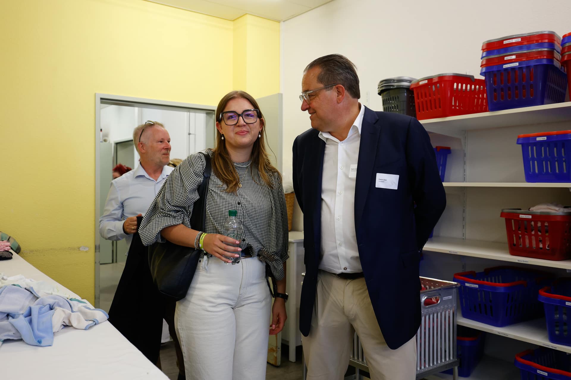 Drei Personen stehen in einem Waschraum, in dem Regale mit roten und blauen Körben stehen; eine junge Frau mit Brille hält eine Wasserflasche in der Hand, während sie mit einem Mann im Anzug spricht, und ein weiterer Mann steht im Hintergrund.