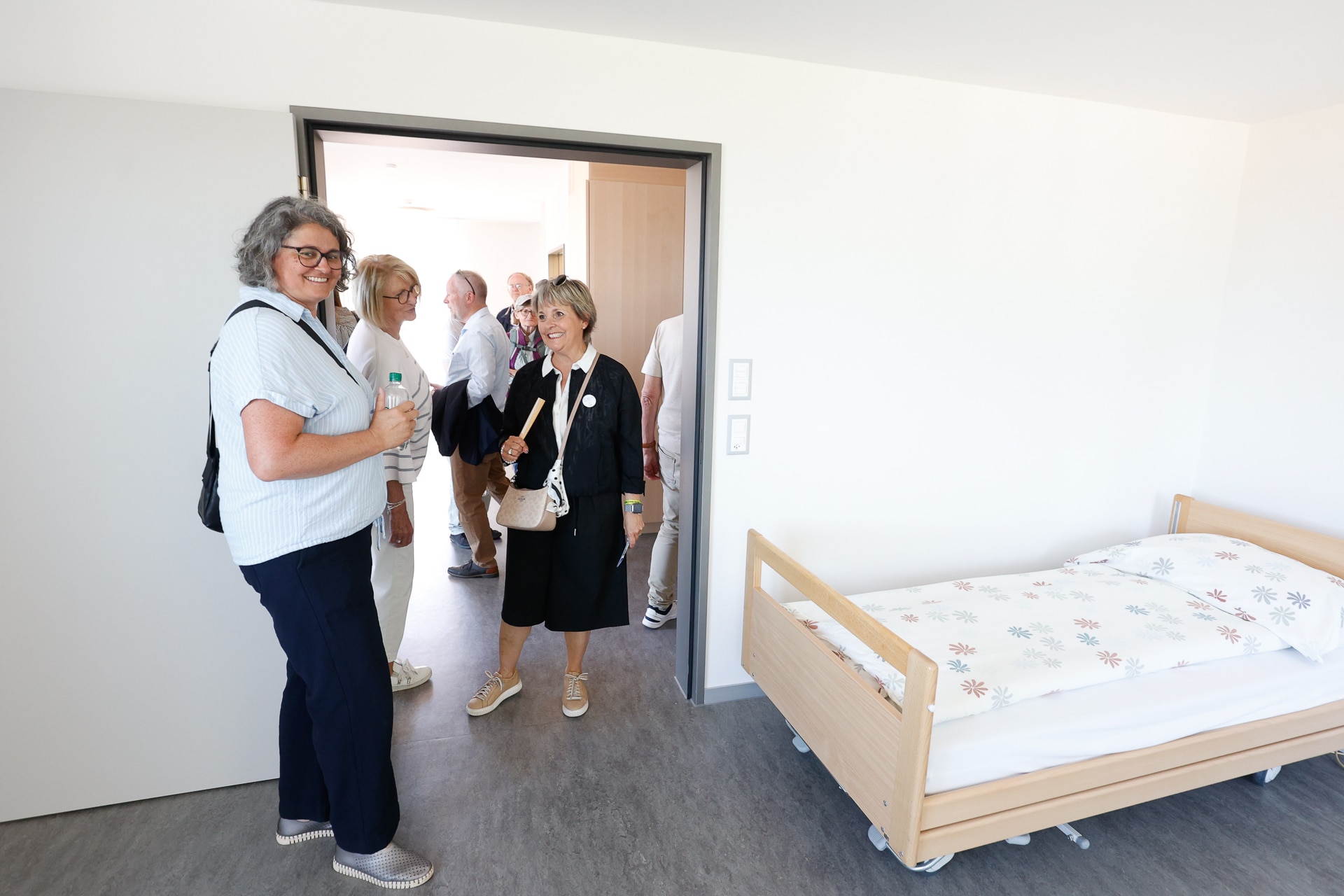 Eine Gruppe Erwachsener steht lächelnd in einem hellen, modernen Zimmer mit einem Einzelbett und gemusterter Bettwäsche. Einige Personen befinden sich im Zimmer, während andere sich in der Tür versammelt haben, was auf eine Führung oder einen Besuch hindeutet.