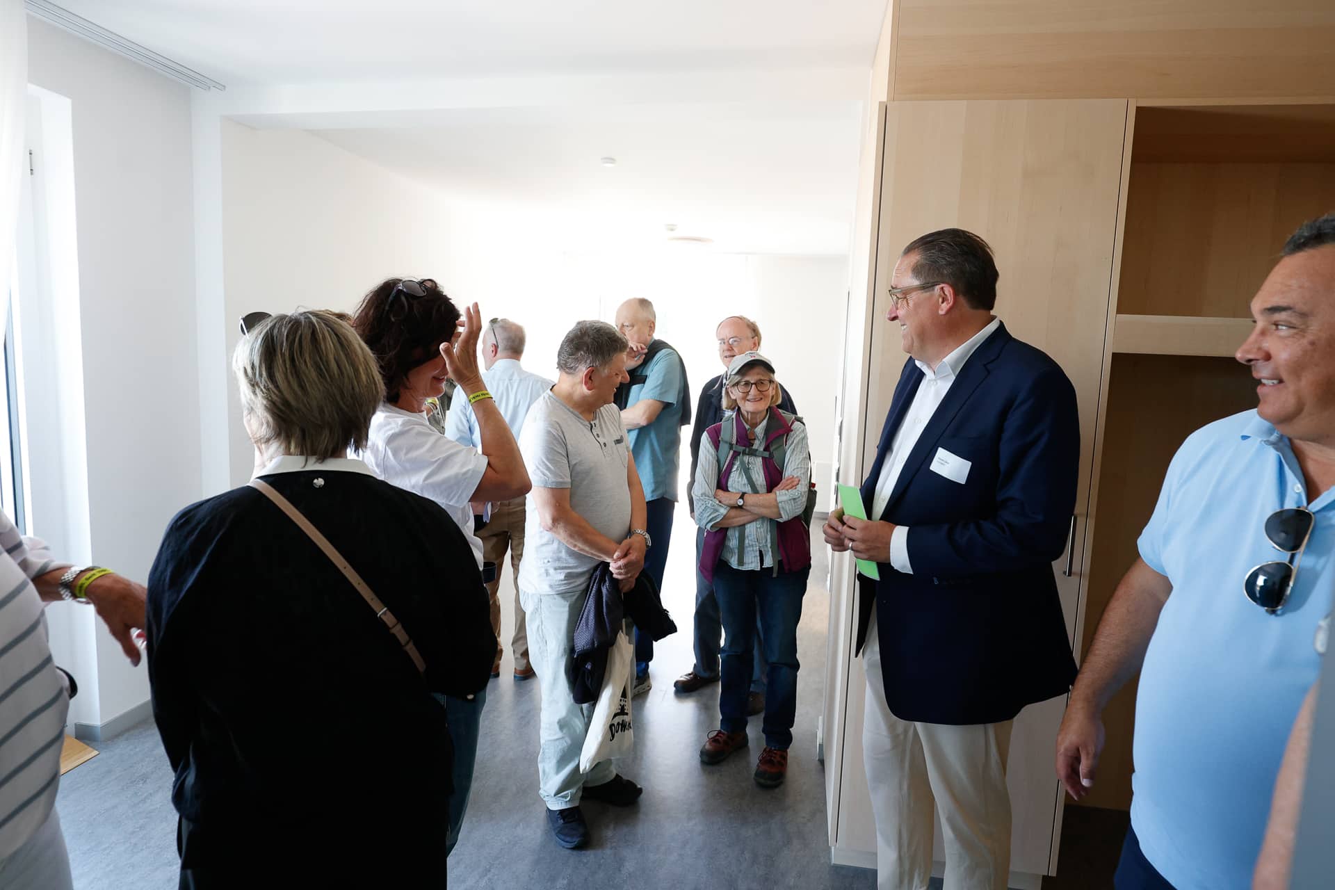 In einem hellen, modernen Raum mit weißen Wänden und Holzschränken steht eine Gruppe von Menschen und unterhält sich. Einige lächeln und tragen Freizeitkleidung, während ein Mann im Anzug neben dem Schrank steht und Unterlagen in der Hand hält.
