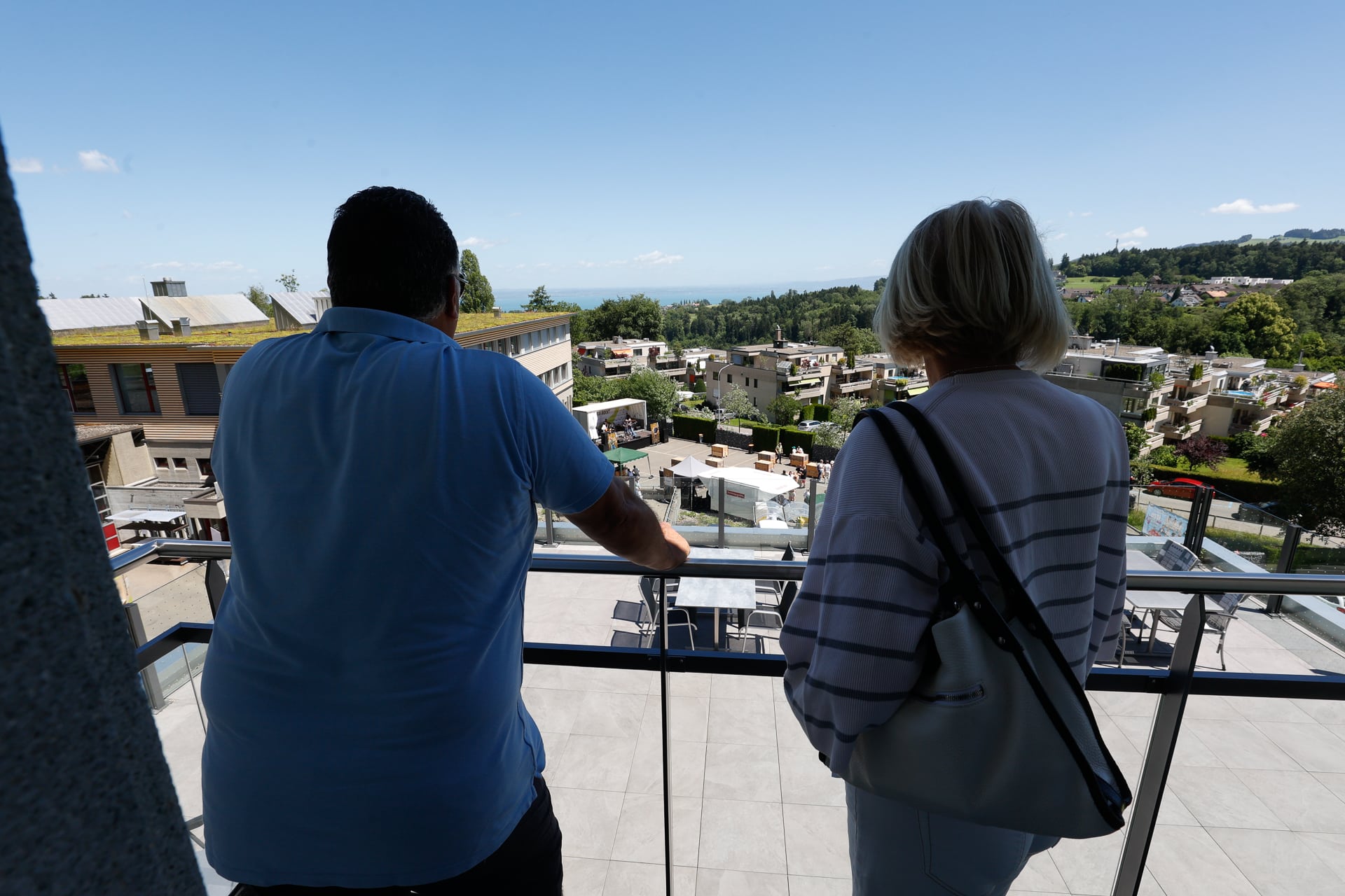 Zwei Menschen stehen auf einem Balkon und blicken auf ein sonniges, modernes Wohngebiet mit viel Grün und Gebäuden in der Ferne unter einem strahlend blauen Himmel.