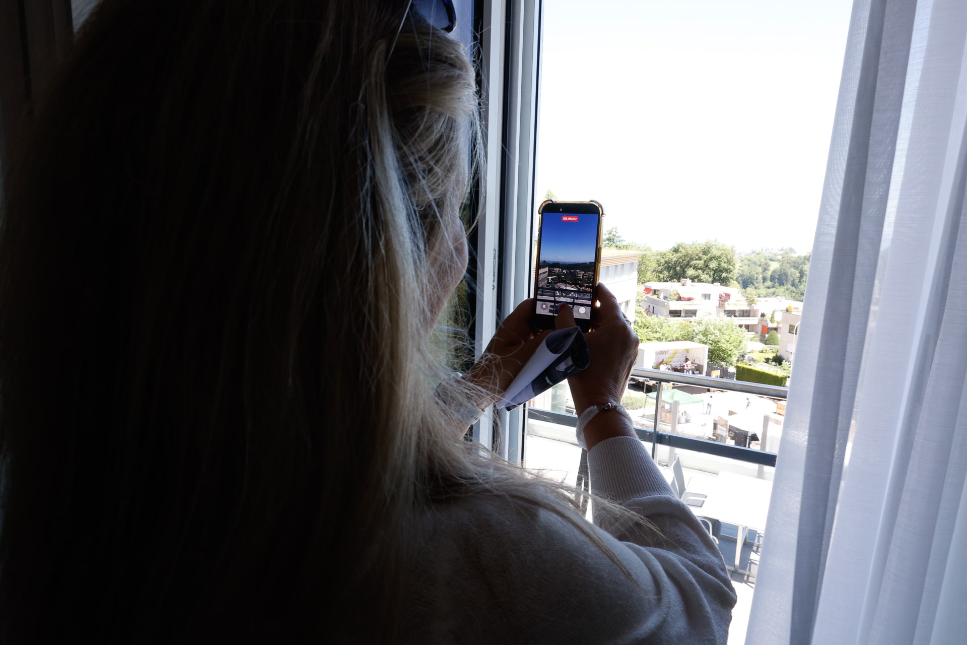 Eine Person mit langen Haaren steht am Fenster, hält ein Handy in der Hand und fotografiert eine sonnige Außenaufnahme, auf der Gebäude, Grünflächen und ein strahlender Himmel zu sehen sind. Weiße Vorhänge rahmen das Fenster ein.