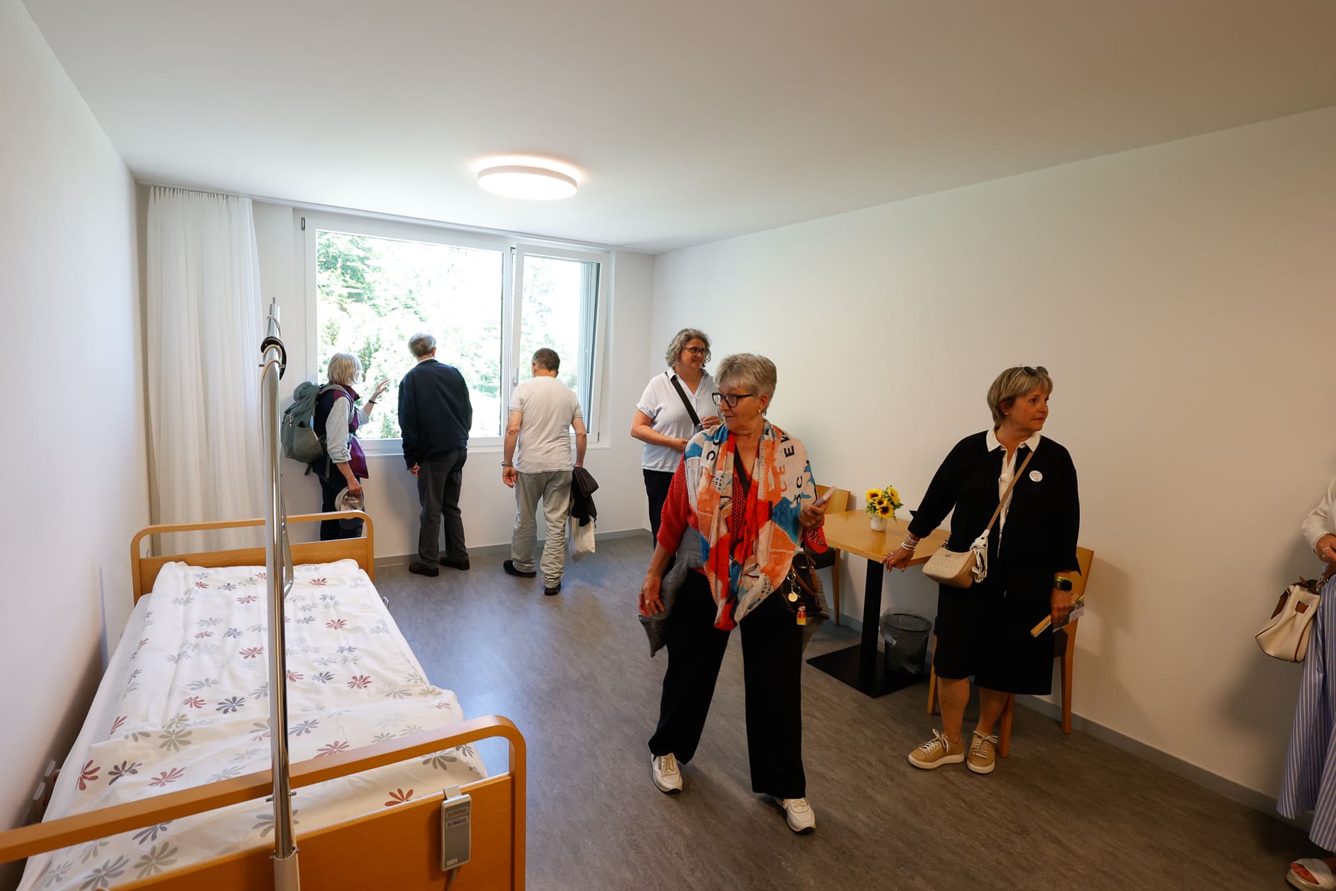 Eine Gruppe von Menschen betrachtet einen schlichten, hellen Raum mit einem einzigen Bett im Krankenhausstil, Holzmöbeln und einem großen Fenster. Einige stehen am Fenster, während andere sich umsehen oder miteinander sprechen.