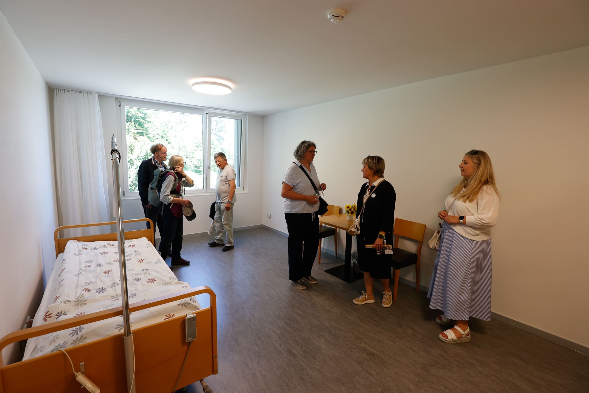 Eine Gruppe von Menschen steht in einem hellen, sparsam eingerichteten Krankenhaus- oder Pflegebereich und unterhält sich. Der Raum ist mit einem Bett, einem kleinen Tisch und einem großen Fenster ausgestattet, durch das Tageslicht hereinfällt.