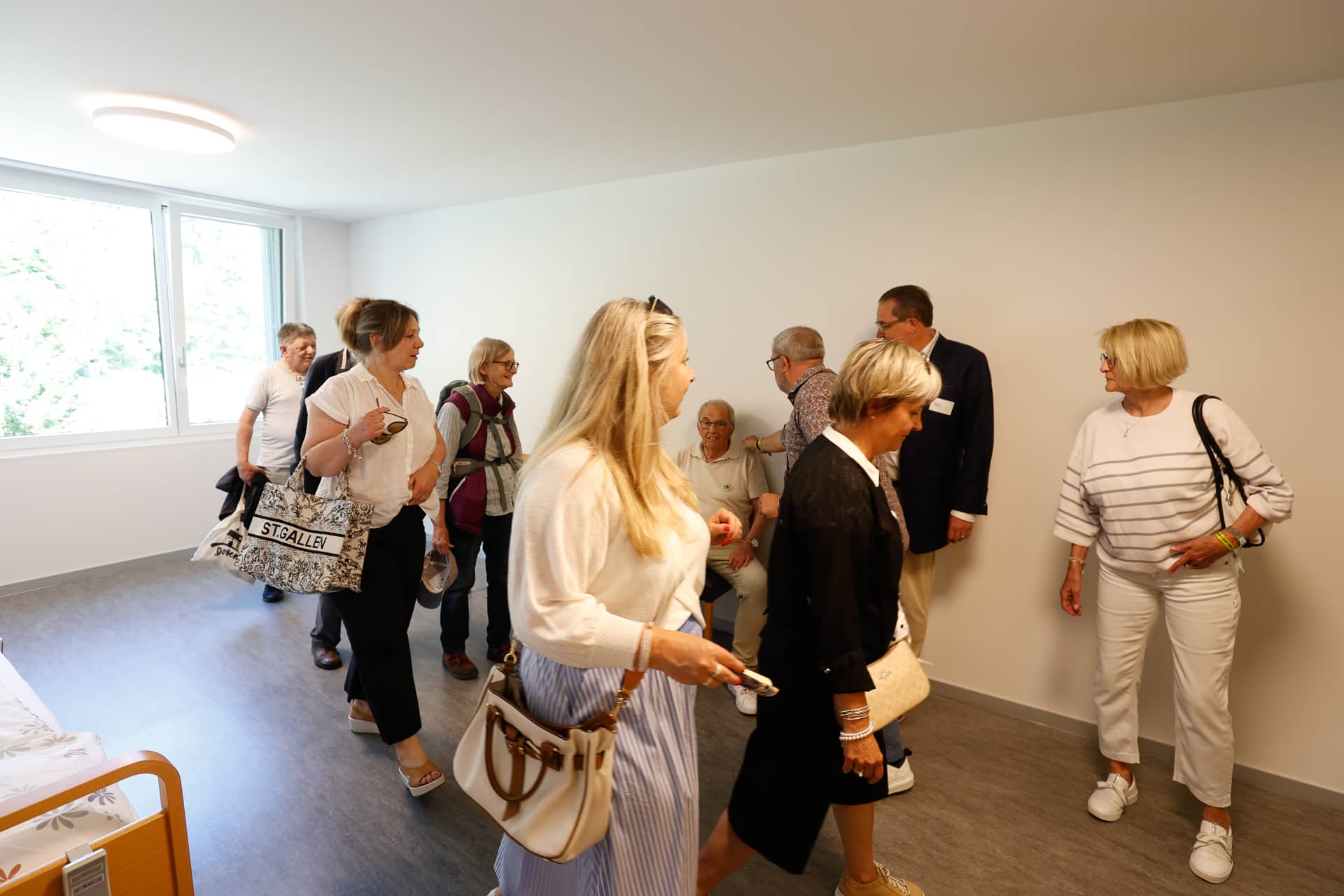 Eine Gruppe von Erwachsenen, überwiegend Frauen, steht in einem hellen, spärlich möblierten Raum mit einem großen Fenster und unterhält sich. Einige lächeln und tauschen sich aus, während ein paar andere im Hintergrund auf Stühlen sitzen.