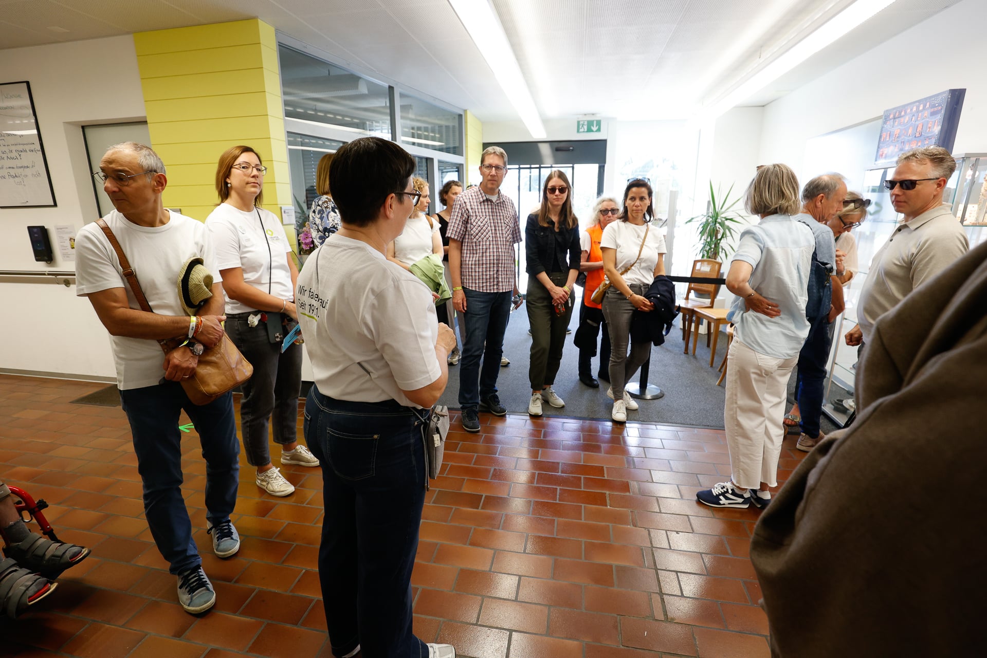Eine Gruppe von Menschen steht in einem hell beleuchteten Flur mit Fliesenboden und großen Fenstern und unterhält sich. Einige tragen Freizeitkleidung und Sonnenbrillen, und ein paar halten Taschen in der Hand.