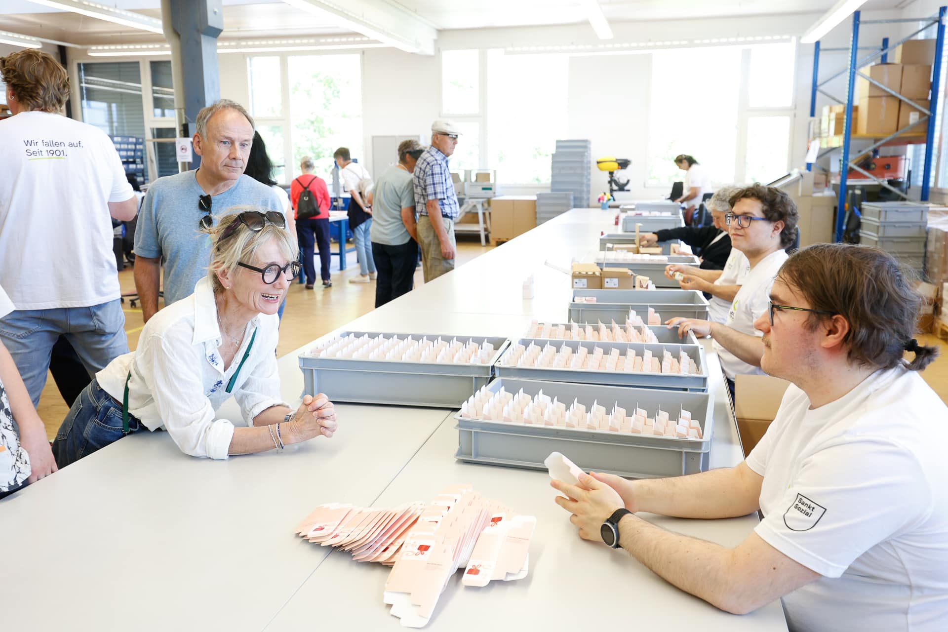 Eine Frau beugt sich über einen Tisch, lächelt und unterhält sich mit einem sitzenden Arbeiter, der in einer hellen, geschäftigen Fabrik oder Werkstatt Produkte zusammenbaut, während andere im Hintergrund zuschauen und miteinander interagieren.