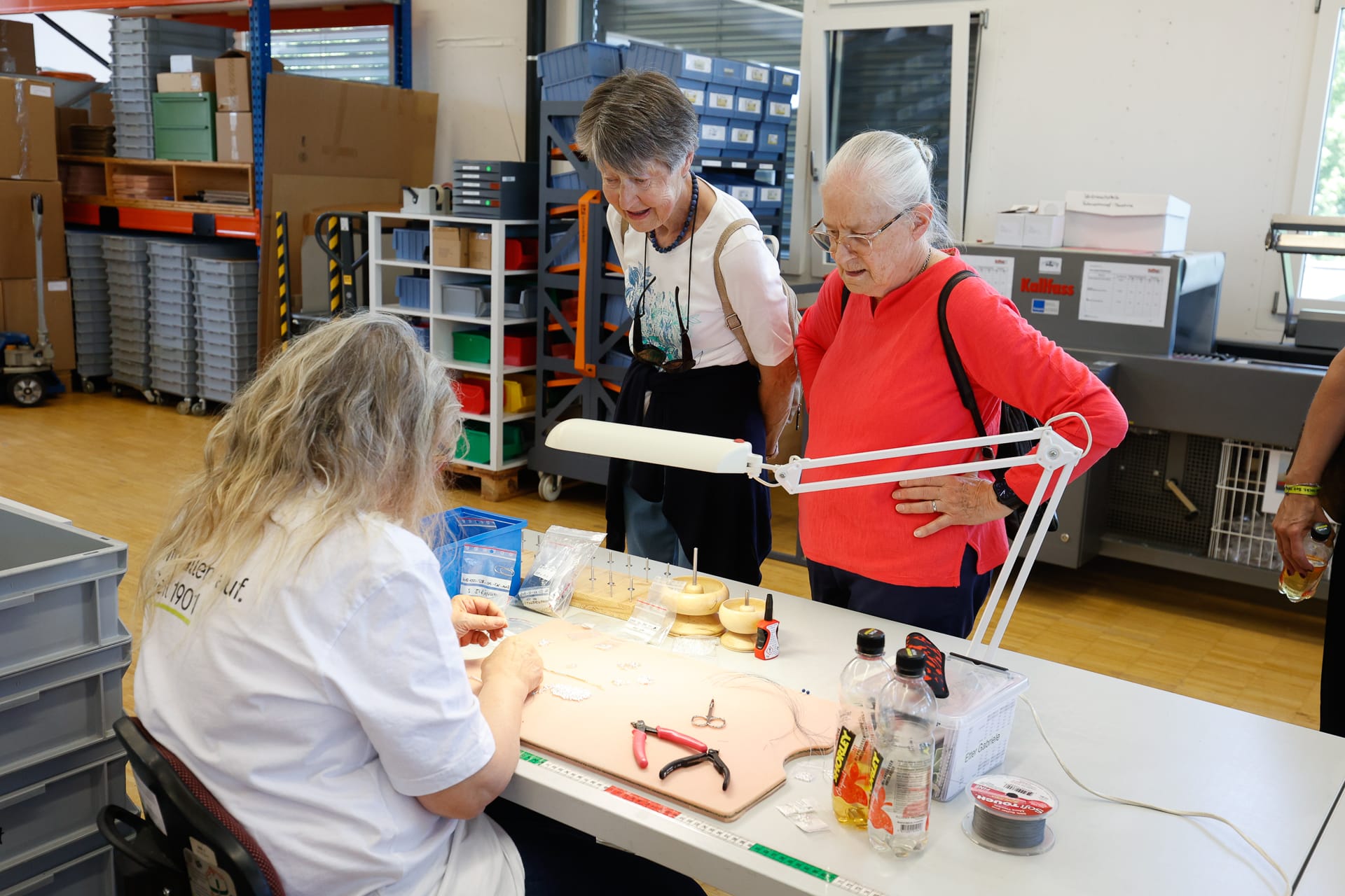 Eine Person sitzt an einem Arbeitstisch und montiert kleine Teile, während zwei ältere Frauen daneben stehen und zusehen. Auf dem Tisch liegen Werkzeuge und Materialien, und die Umgebung scheint eine Werkstatt oder Fabrik zu sein, da im Hintergrund Regale und Kisten zu sehen sind.