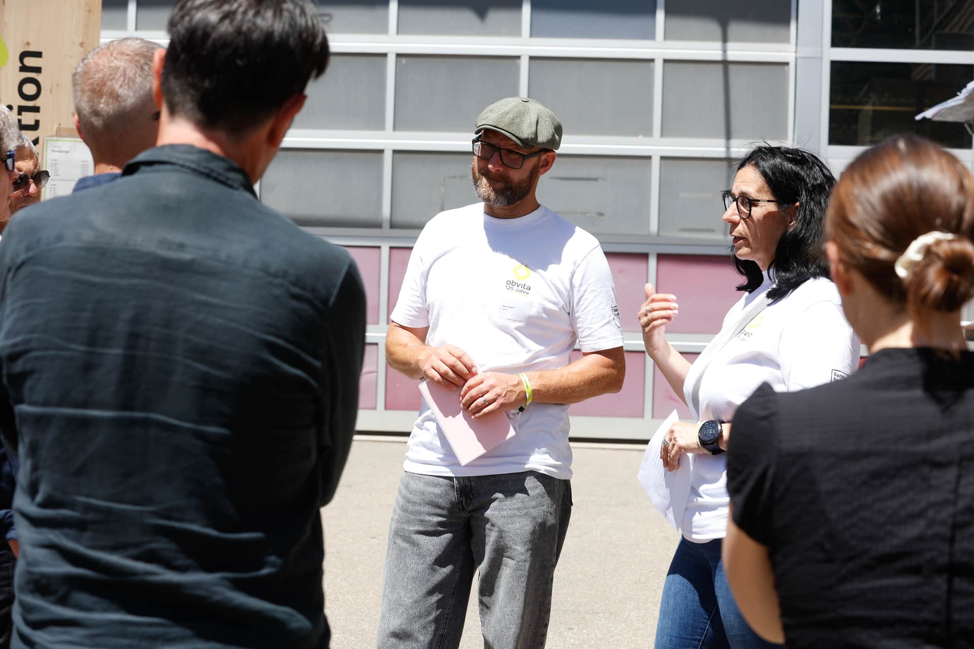 Ein Mann und eine Frau, beide in weißen T-Shirts, sprechen im Freien zu einer Gruppe von Menschen. Der Mann hält Papiere in der Hand, und die Frau untermalt ihre Worte mit Gesten. Die Gruppe steht in der Nähe eines Gebäudes mit großen Fenstern.