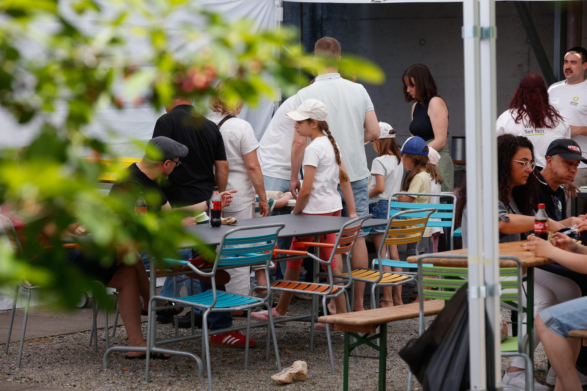 Die Menschen versammeln sich im Freien an bunten Tischen und Stühlen, essen, trinken und unterhalten sich. Einige Erwachsene und Kinder stehen oder sitzen, während belaubte Äste den Blick im Vordergrund teilweise einrahmen. Die Atmosphäre wirkt entspannt und gesellig.