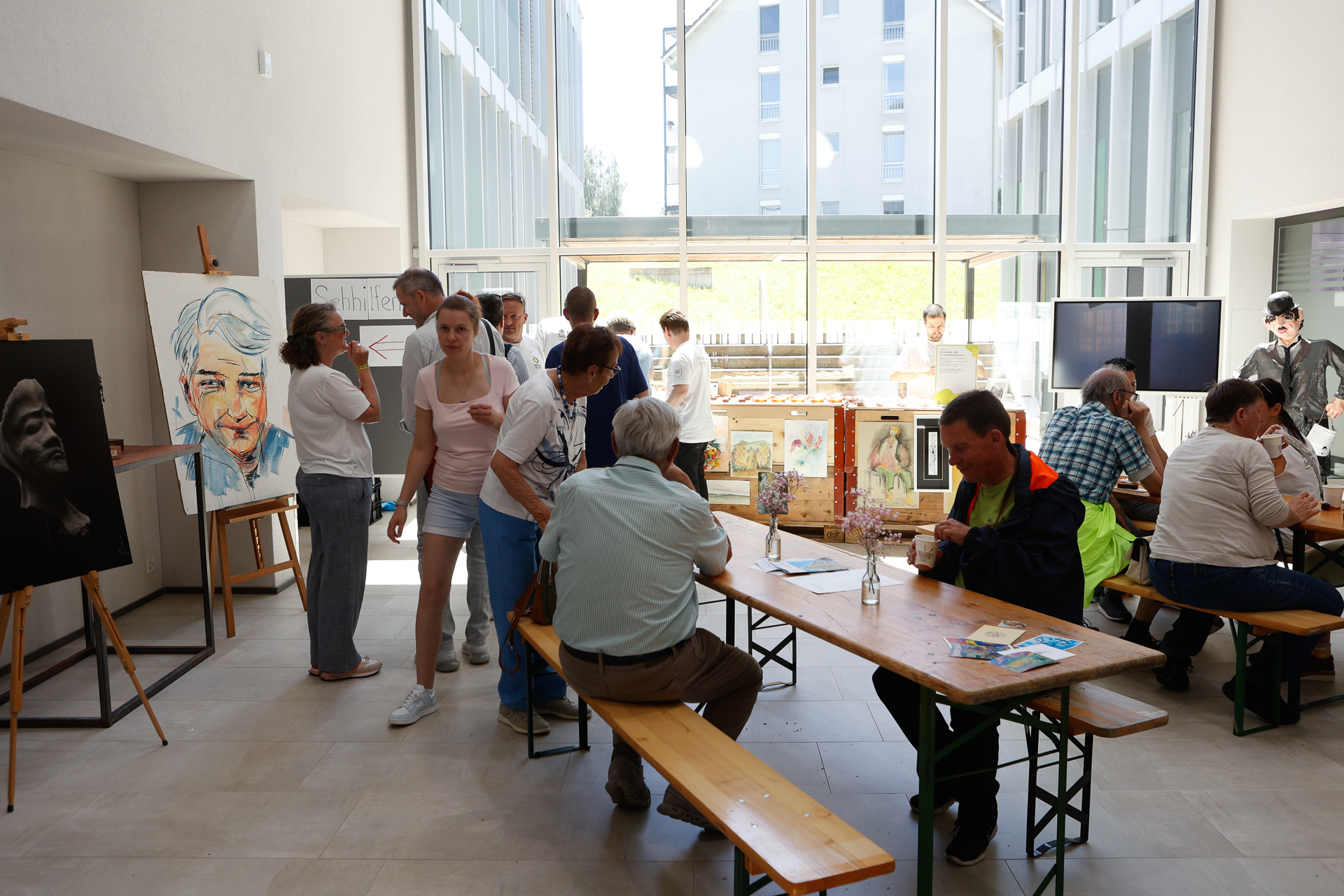 Die Menschen versammeln sich in einem hellen Kunstatelier mit großen Fenstern, betrachten die auf Staffeleien ausgestellten Kunstwerke und diskutieren darüber. Einige sitzen an langen Holztischen, während andere umhergehen, was eine lebhafte und gesellige Atmosphäre schafft.
