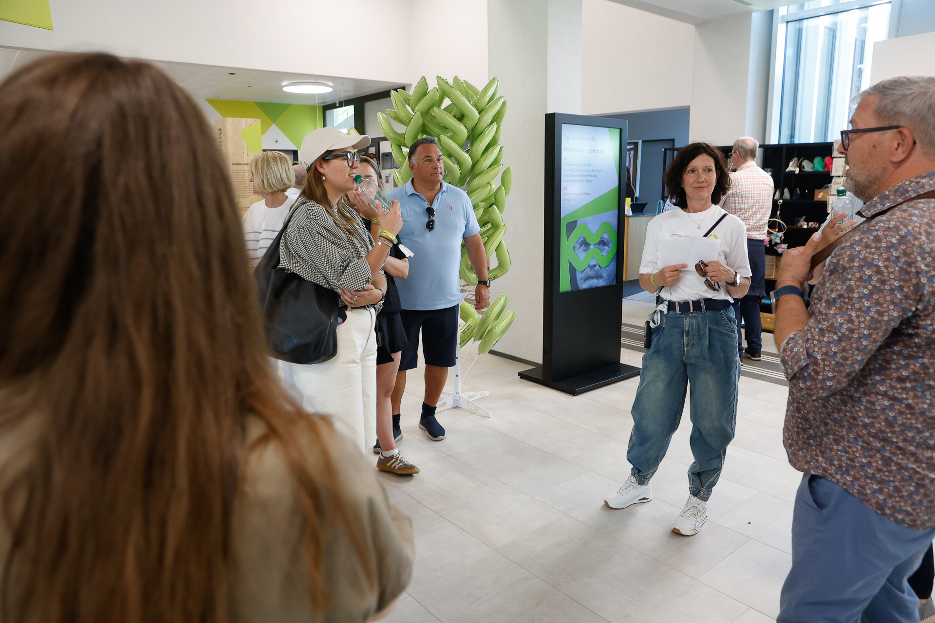Eine Gruppe von Menschen steht in einem Innenraum in der Nähe eines digitalen Displays und einer grünen Pflanzendekoration. Eine Frau hält Papiere in der Hand, während die anderen sich unterhalten und zuhören. Der Schauplatz scheint eine moderne Lobby oder ein Veranstaltungsraum zu sein.