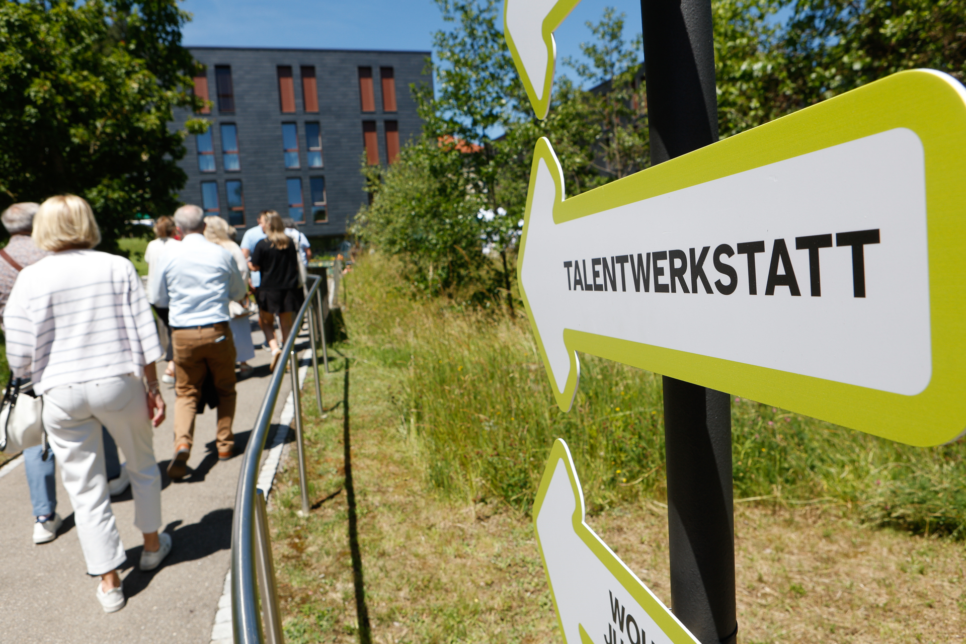 Eine Gruppe von Menschen geht auf einem Weg neben einer Grünfläche entlang in Richtung eines modernen Gebäudes. Im Vordergrund ist ein gelb-weißes Schild mit der Aufschrift „TALENTWERKSTATT“ zu sehen.