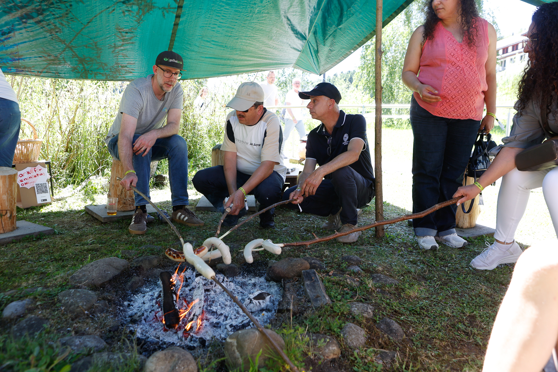 Vier Menschen sitzen unter einem Vordach und rösten Brotteig auf Stöcken über einem Lagerfeuer, während andere an einem sonnigen Tag in der Nähe stehen. Die Szene spielt sich im Freien ab, im Hintergrund sind Gras und Bäume zu sehen.