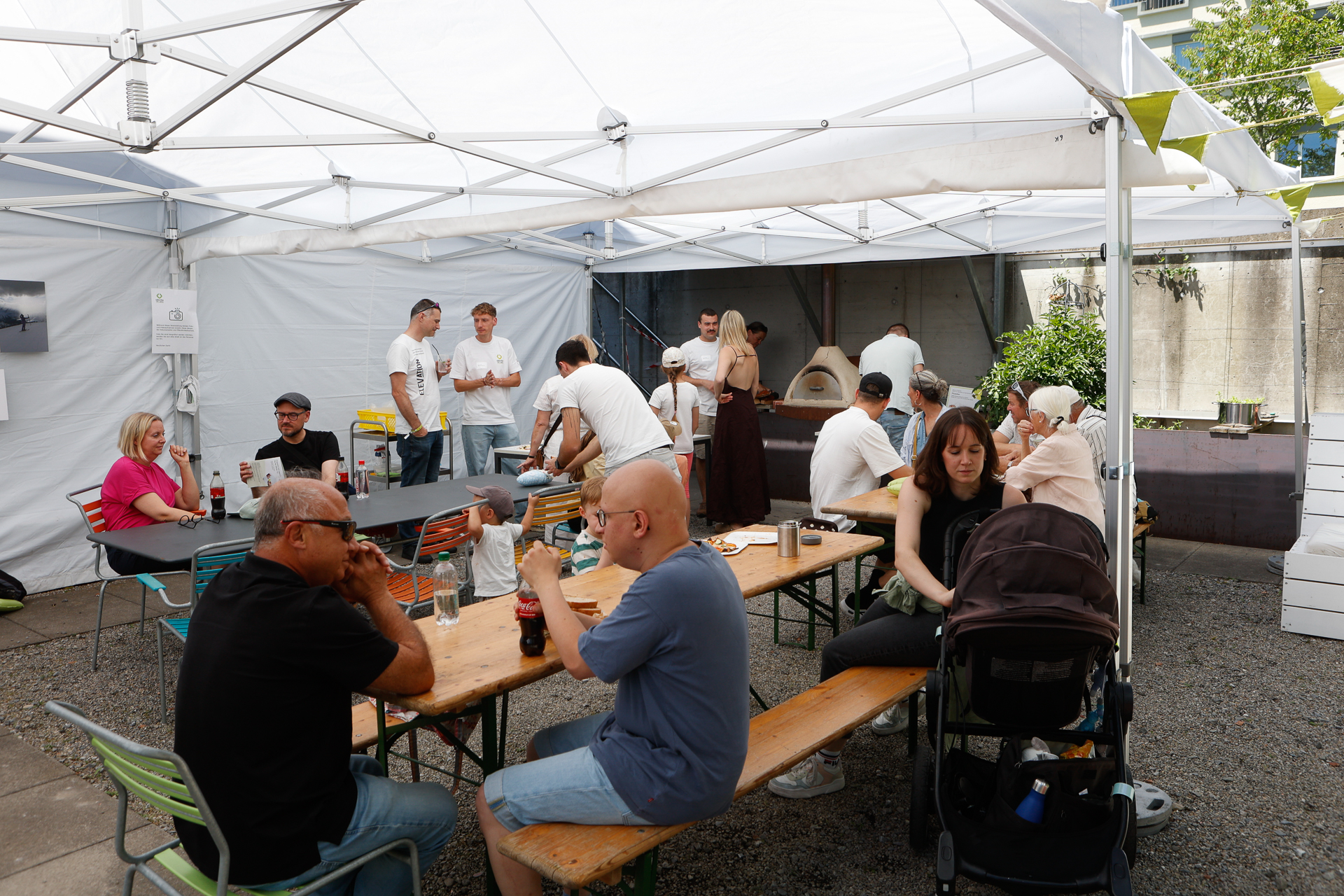 Die Menschen versammeln sich unter einem weißen Festzelt, sitzen an Tischen, unterhalten sich und essen. Einige stehen im Hintergrund in der Nähe eines Pizzaofens. Es sind sowohl Erwachsene als auch Kinder dabei, und die Szene wirkt ungezwungen und gesellig.