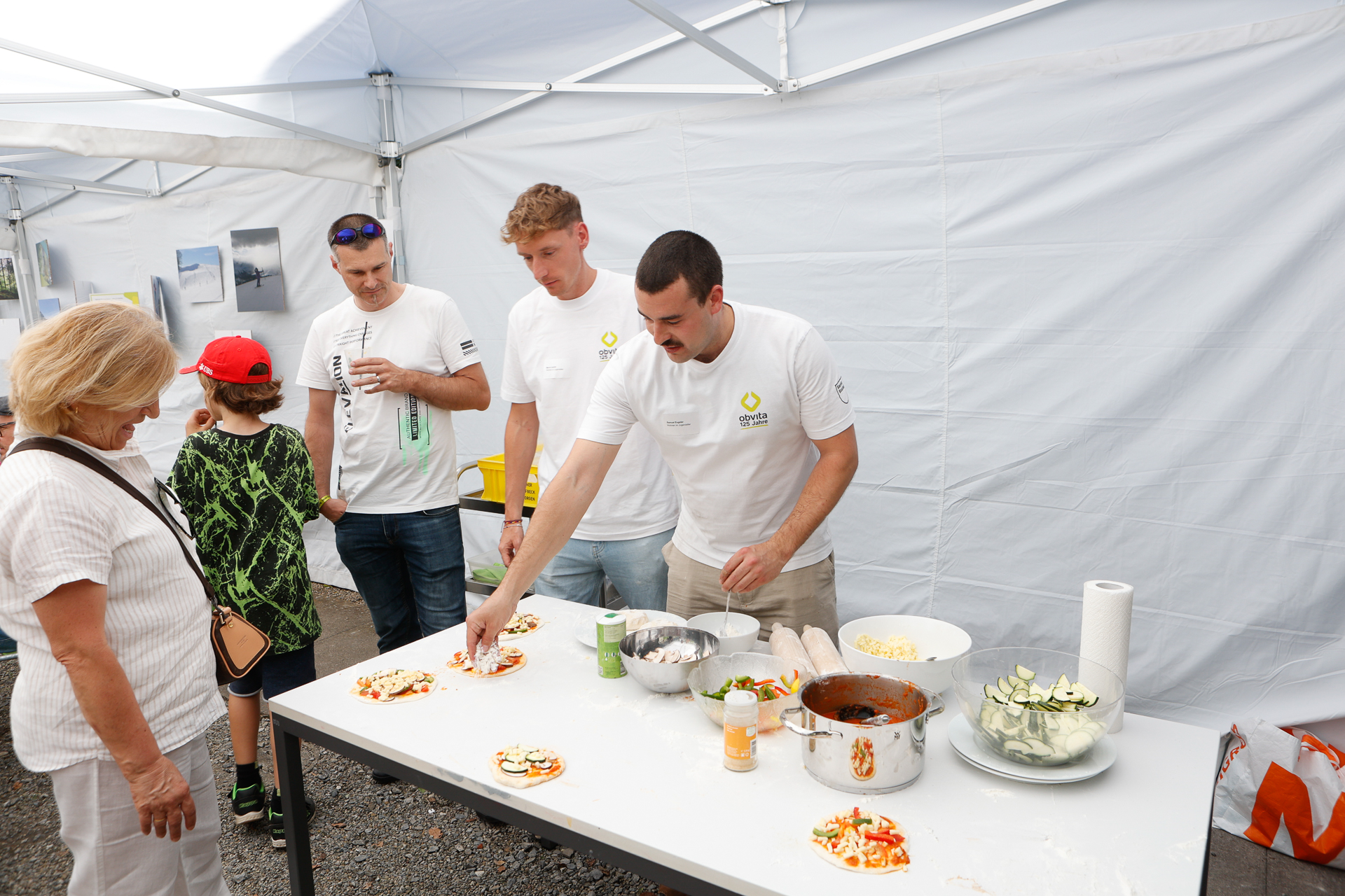 Drei Männer in weißen Hemden bereiten an einem Tisch unter einem Zelt Mini-Pizzen zu, während eine Frau und ein Kind zuschauen. Auf dem Tisch liegen verschiedene Pizzabeläge und Küchenutensilien ausgebreitet.