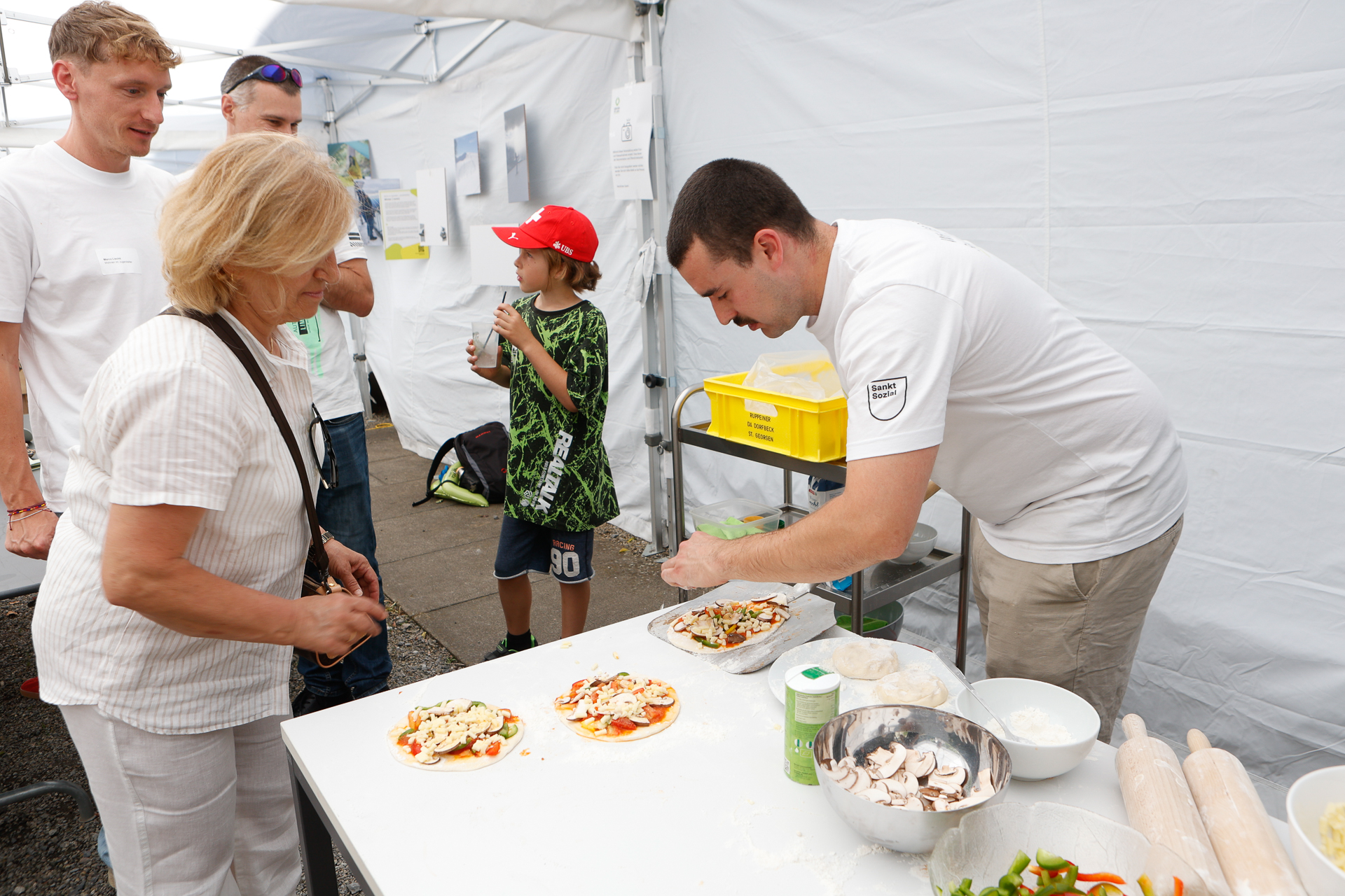 Ein Mann bereitet in einem weißen Zelt bei einer Veranstaltung im Freien auf einem mit Zutaten bedeckten Tisch Pizzen zu, während eine Frau und andere ihm dabei zusehen. Ein Kind mit einer roten Mütze steht daneben, und auf dem Tisch liegen verschiedene Gemüsesorten und Küchenutensilien.