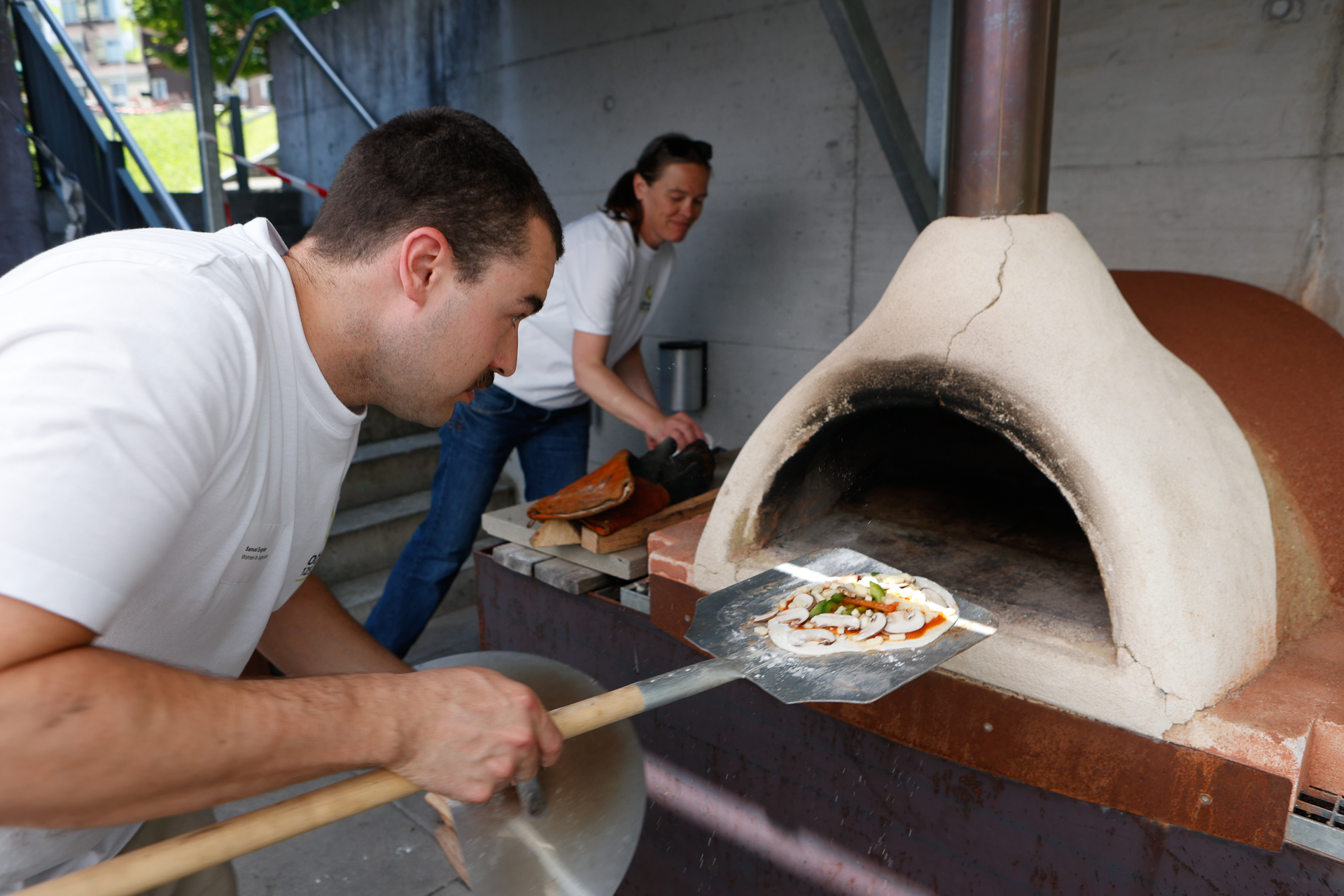 Ein Mann schiebt eine rohe Pizza mit einer Metallschaufel in einen Holzofen, während eine Frau im Hintergrund auf einer Arbeitsplatte Essen zubereitet. Beide tragen weiße Hemden und arbeiten im Freien.