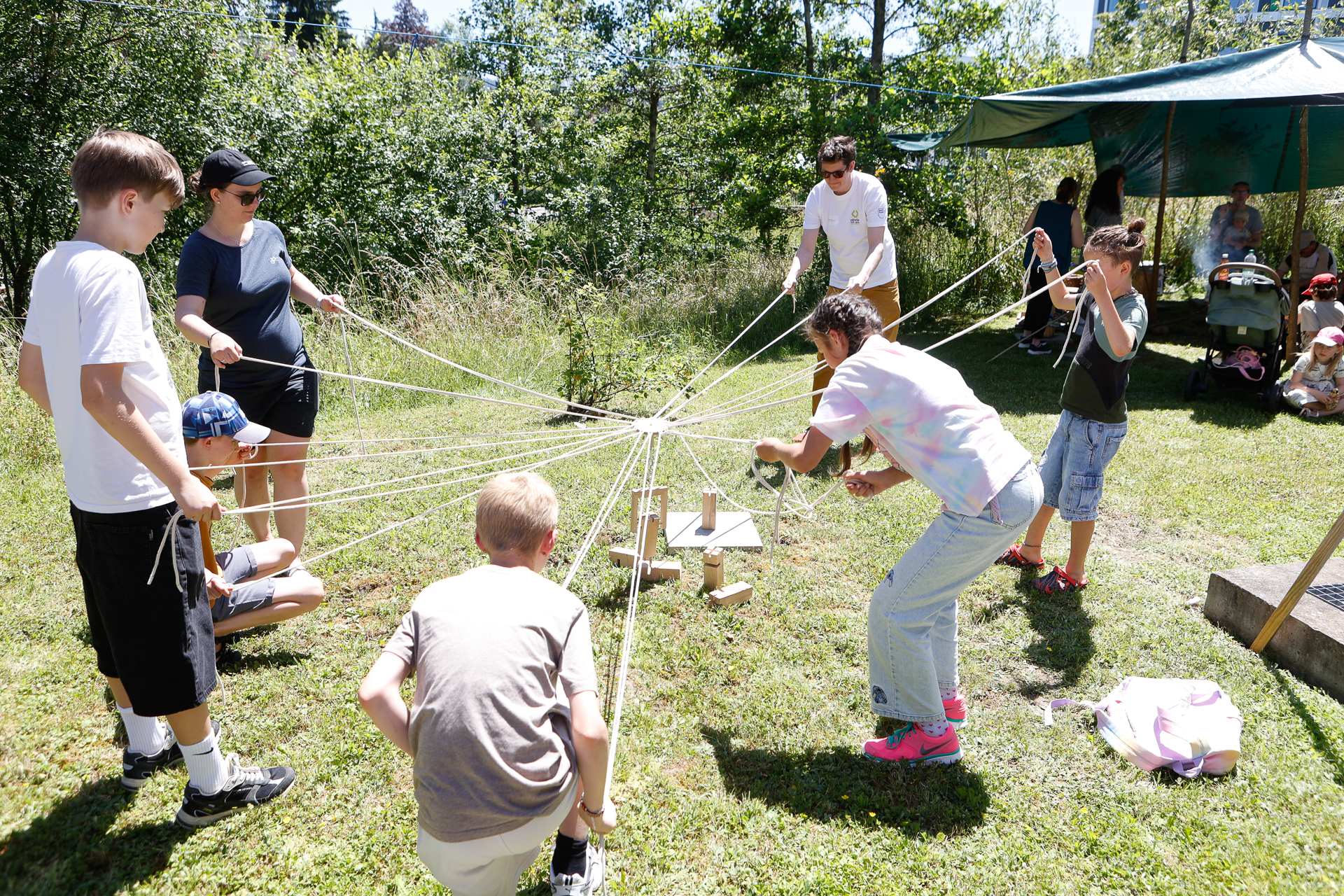 Eine Gruppe von Kindern und zwei Erwachsene steht auf einer Wiese, jeder hält eine Schnur in der Hand, die mit einer zentralen Konstruktion verbunden ist, und nimmt bei sonnigem Wetter gemeinsam an einer Teambildungsübung teil.