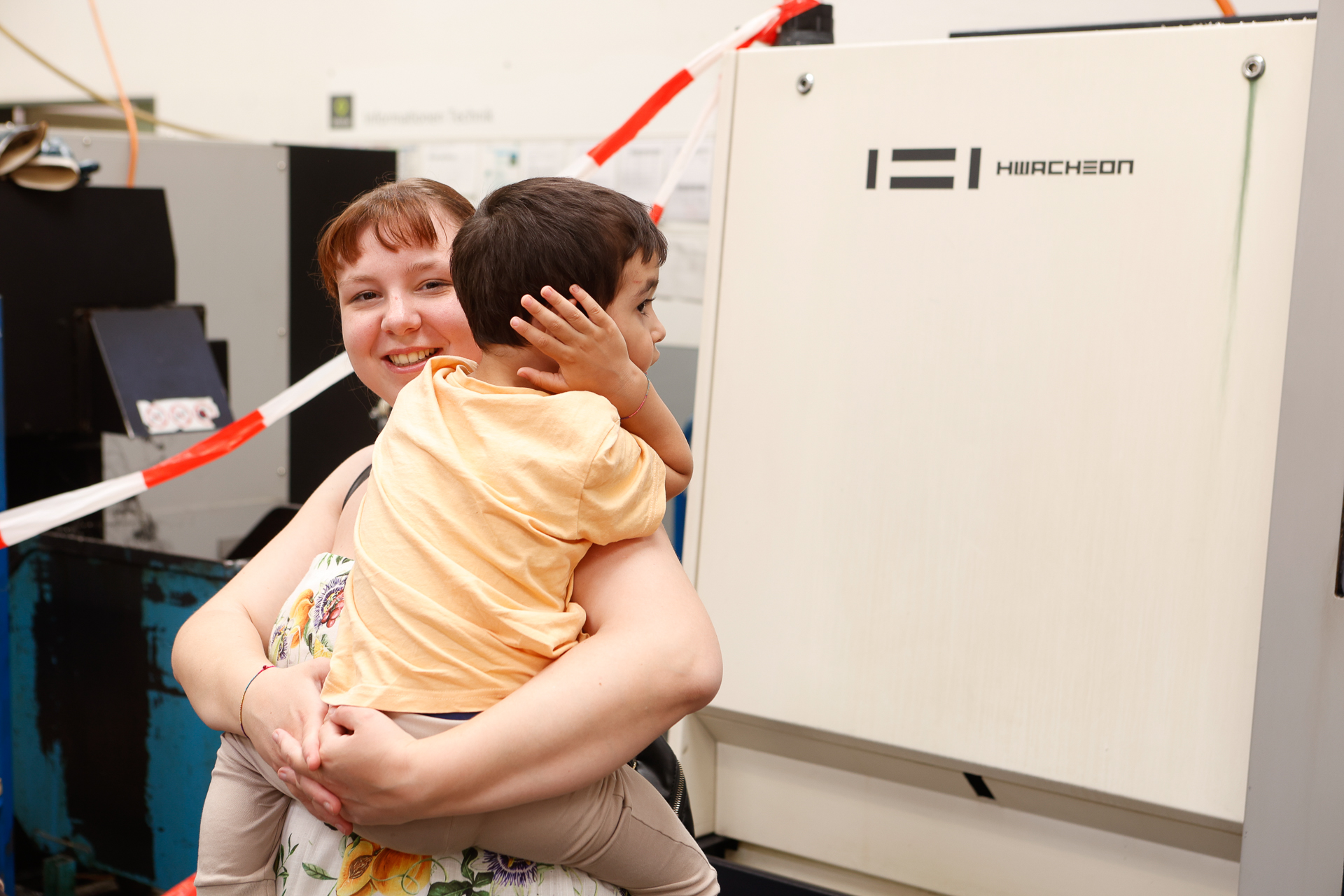 Eine lächelnde Frau hält einen kleinen Jungen im Arm, der sich die Ohren zuhält, und steht vor einer großen Industriemaschine mit der Aufschrift „HIIRCHON“. Im Hintergrund ist rot-weiß gestreiftes Klebeband zu sehen.